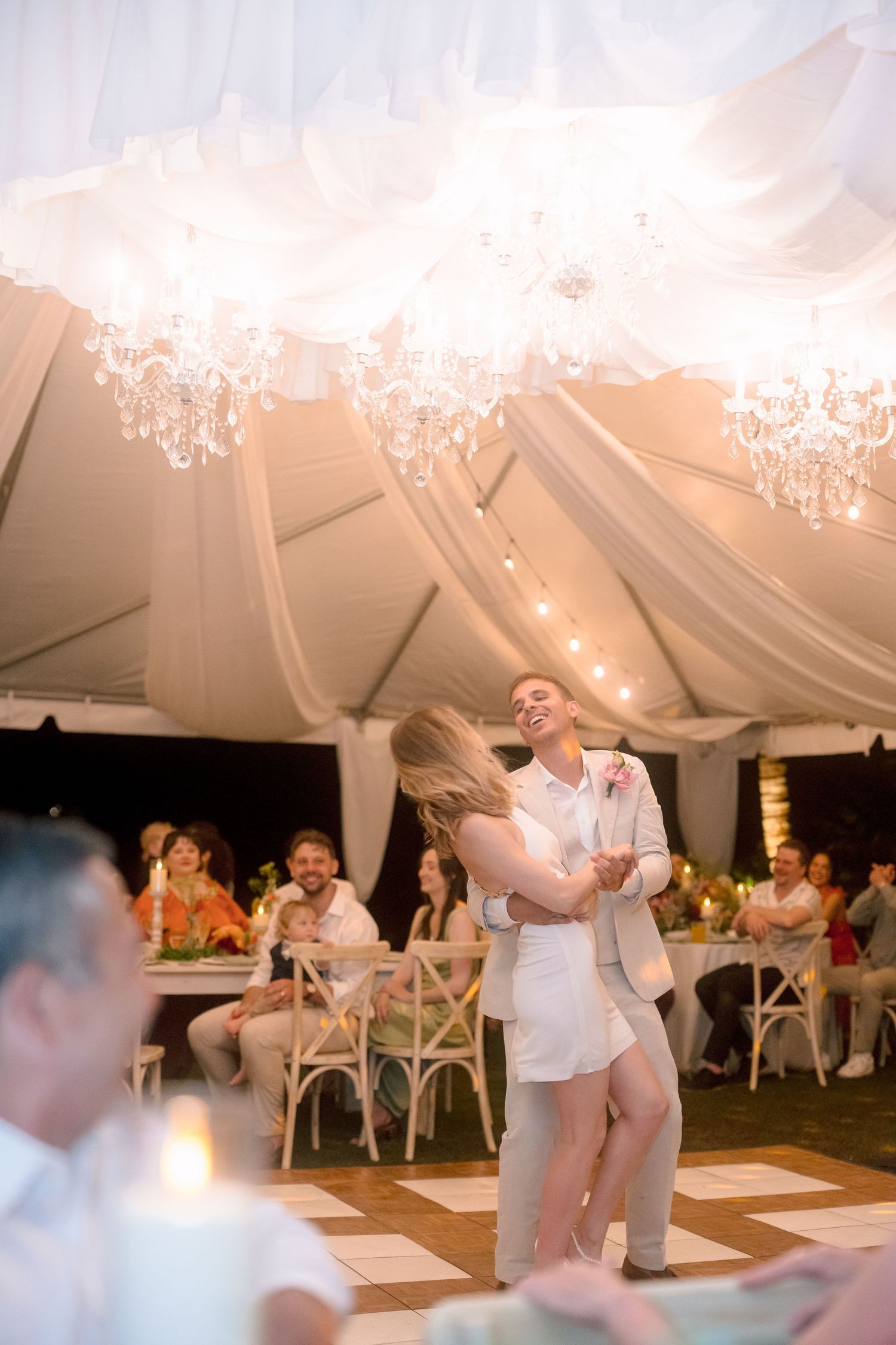 Bride and groom sharing joyful first dance under crystal chandeliers in elegant draped wedding reception tent.