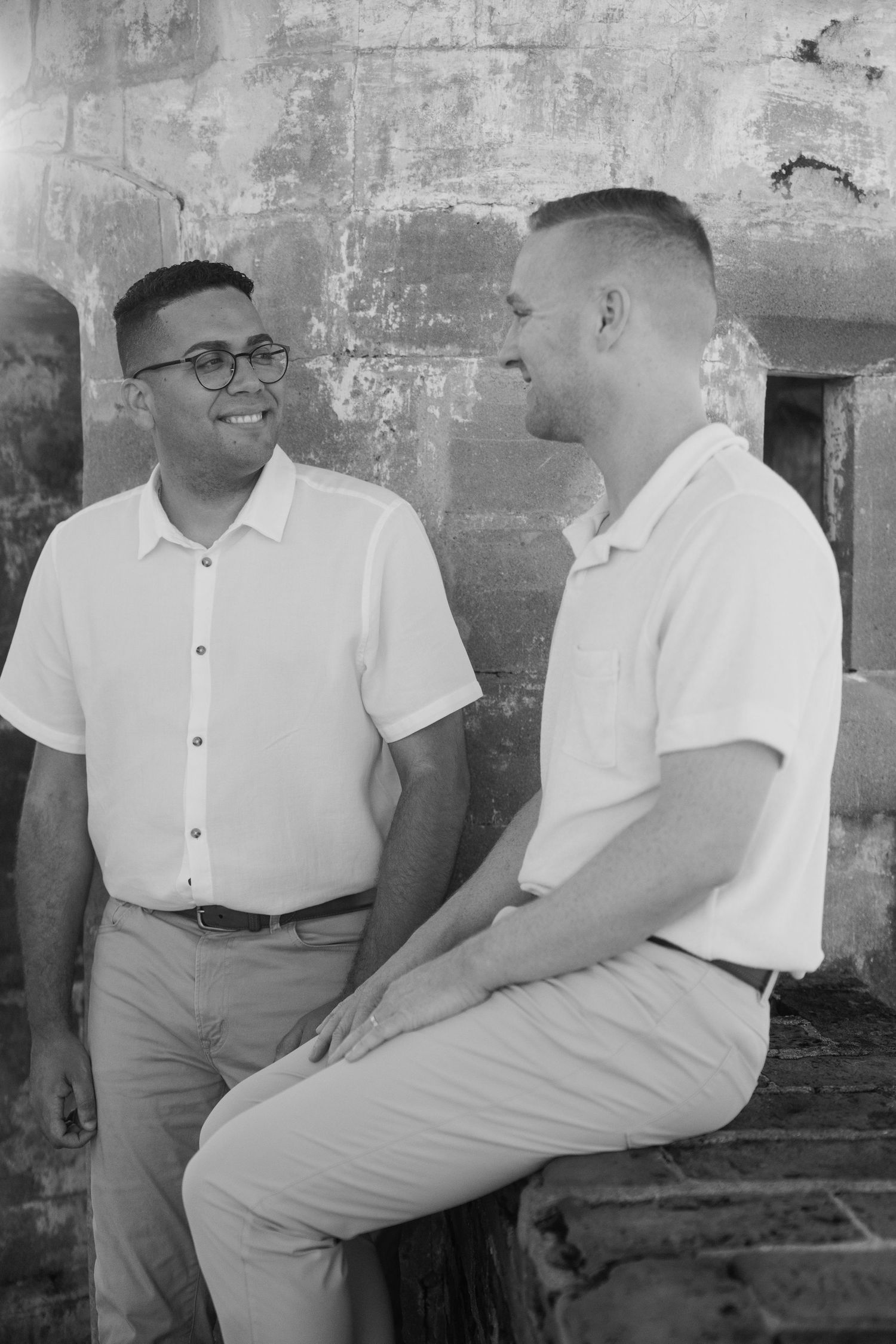 Two men in white shirts share a smile while posing against a rustic stone wall in a black and white photo.