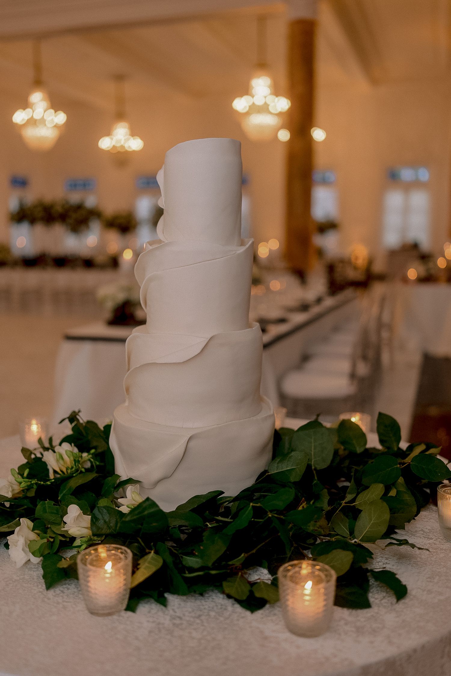 Elegant four-tier white wedding cake with ruffled fondant, surrounded by greenery and candles in a warmly lit reception hall.