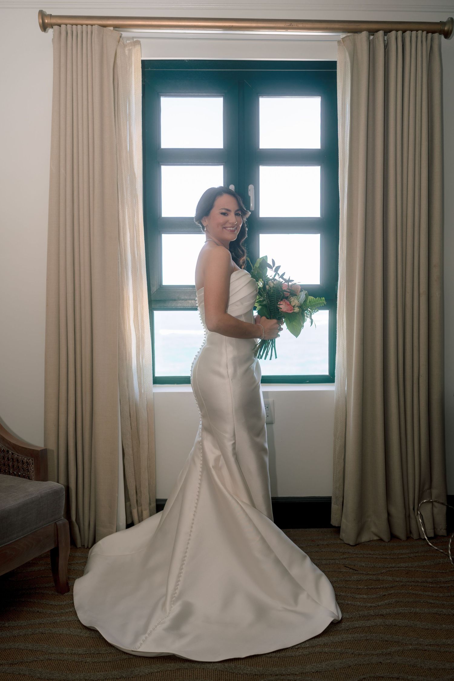 Bride in white mermaid gown holding bouquet stands before window with ocean view on her wedding day.