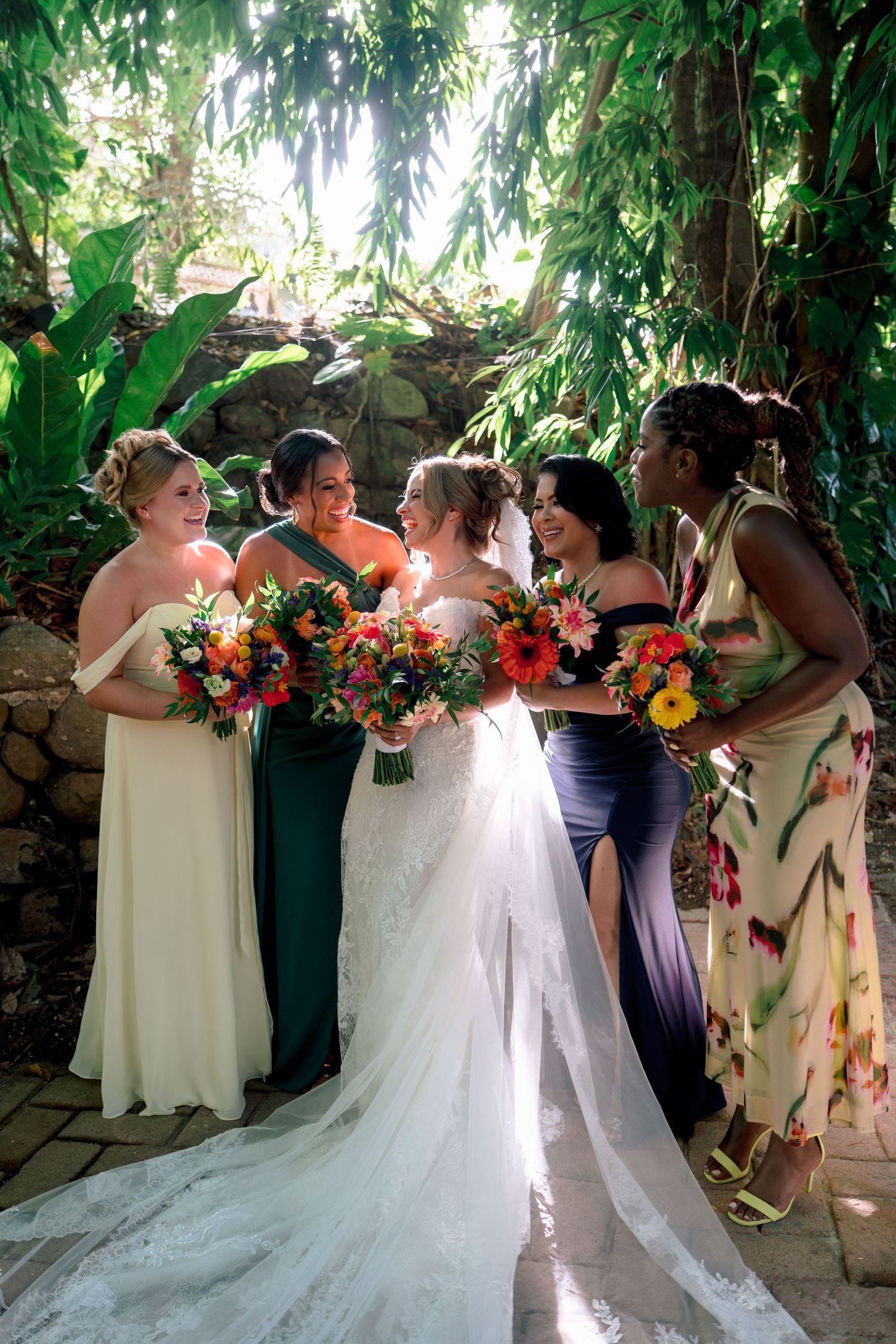 Bride and bridesmaids in colorful gowns holding vibrant bouquets, laughing together in a lush tropical garden setting.