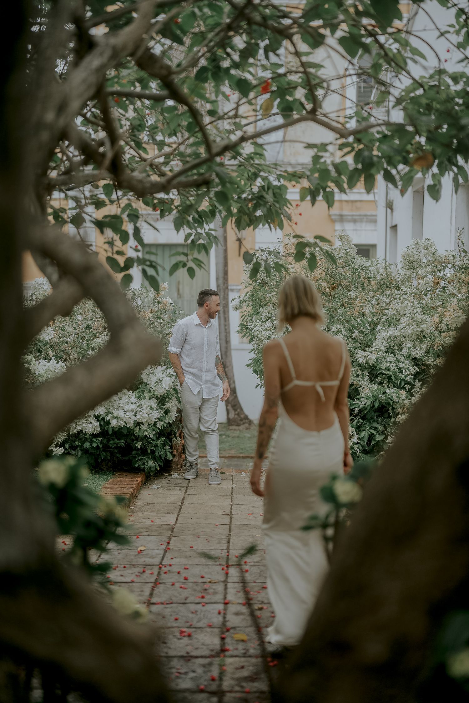 Couple in white wedding attire walking through a lush garden pathway framed by trees and flowering shrubs.