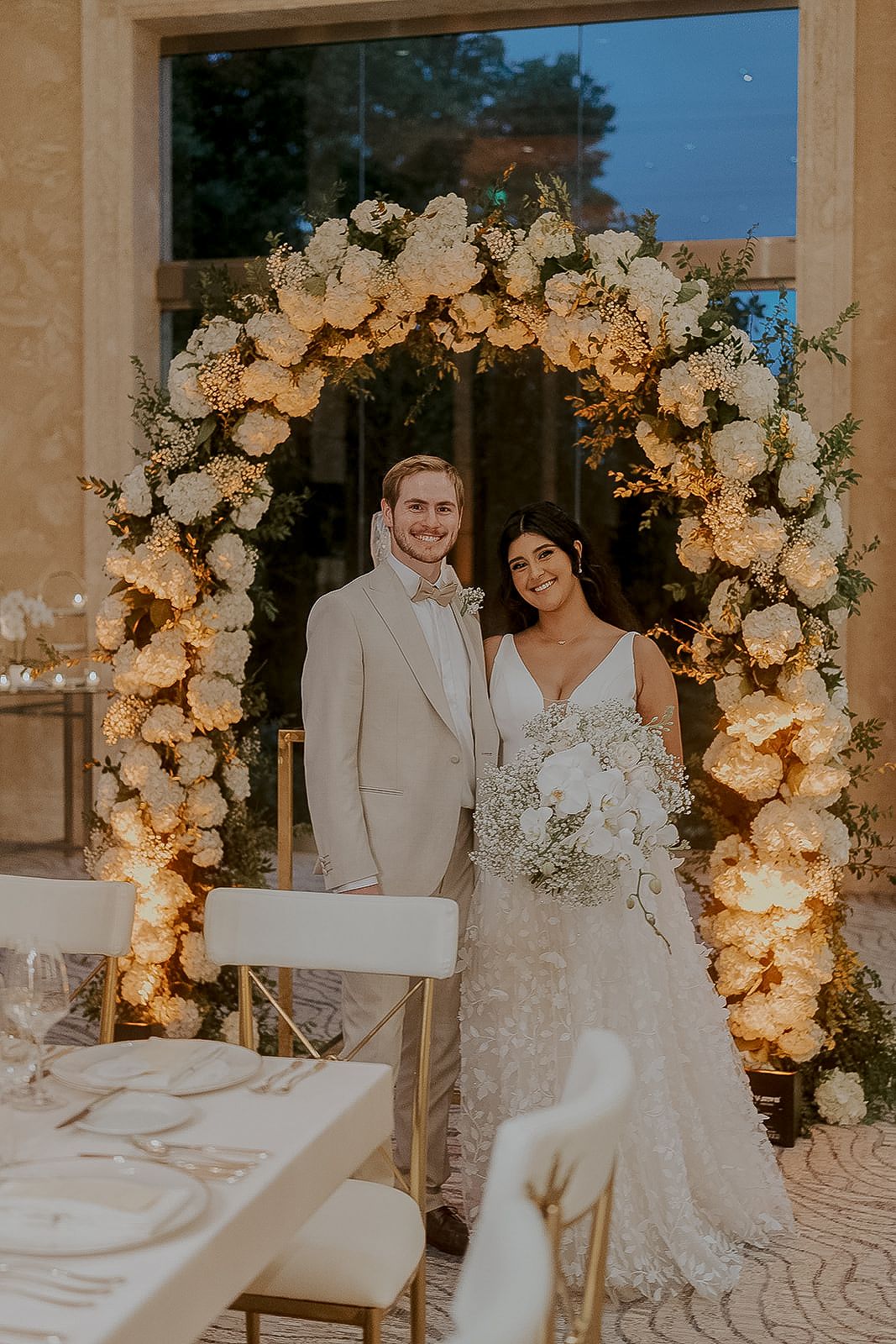 Smiling couple posing under illuminated white floral arch at elegant wedding reception venue.