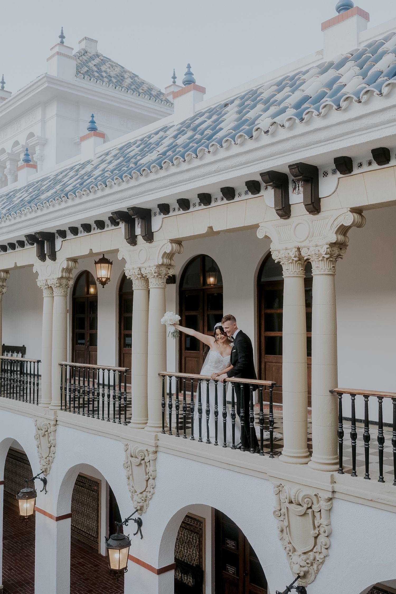 Wedding couple embracing on ornate white Spanish colonial building balcony with arched columns.