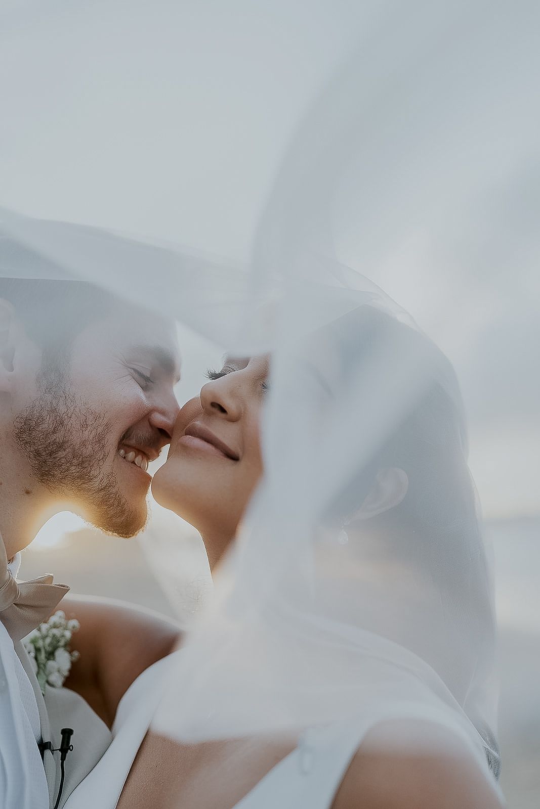 Bride and groom share a tender nose-to-nose moment beneath a billowing white wedding veil.