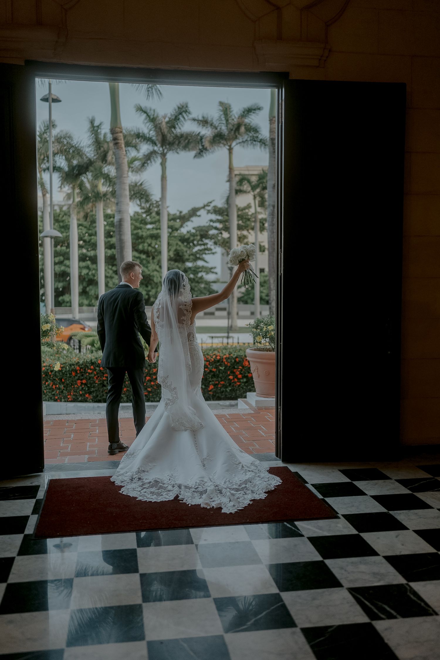 Bride and groom silhouetted in doorway with palm trees and tropical wedding venue in background.