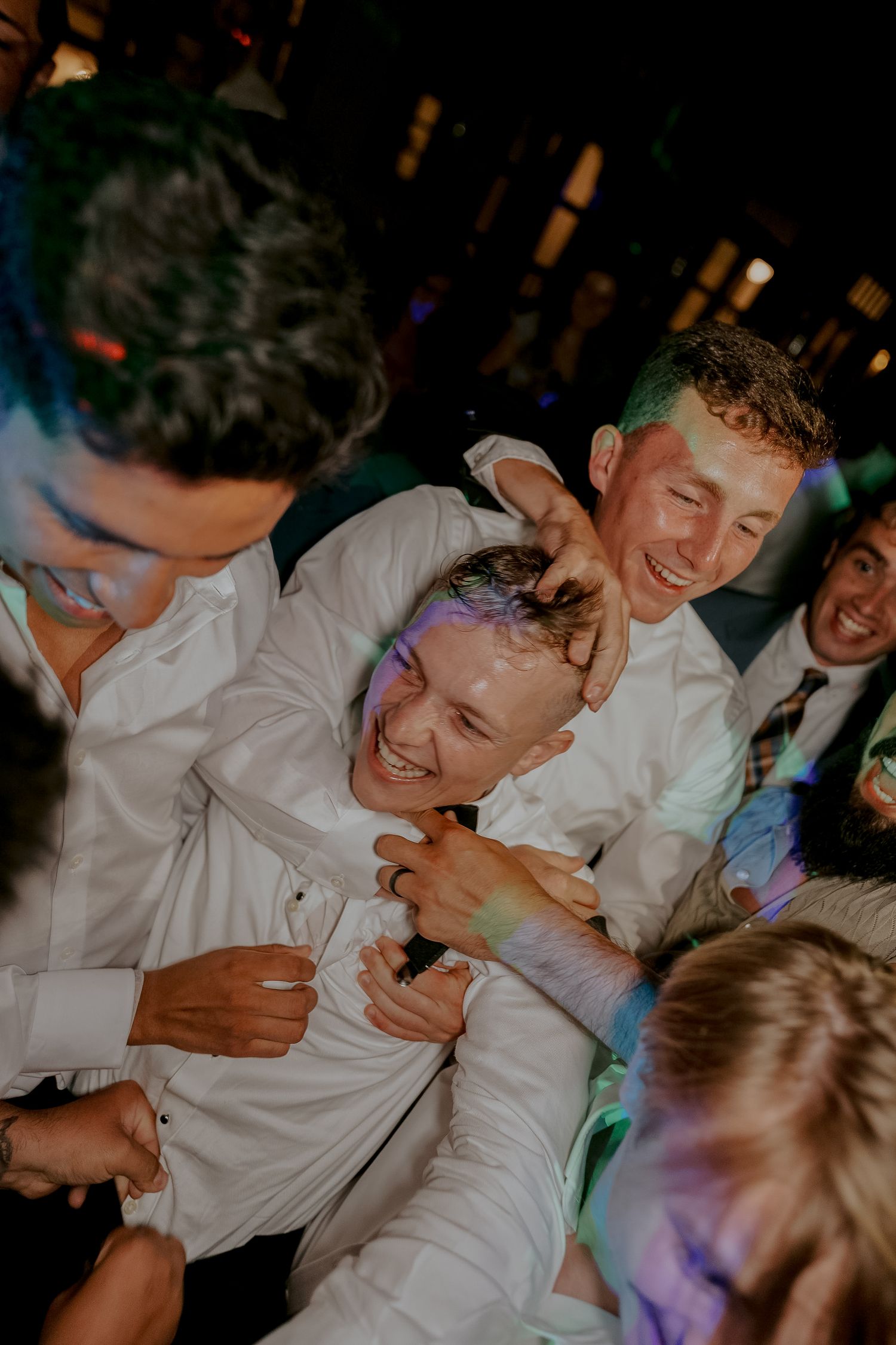 Energetic wedding guests laughing and dancing together under colorful lights at lively wedding reception party.