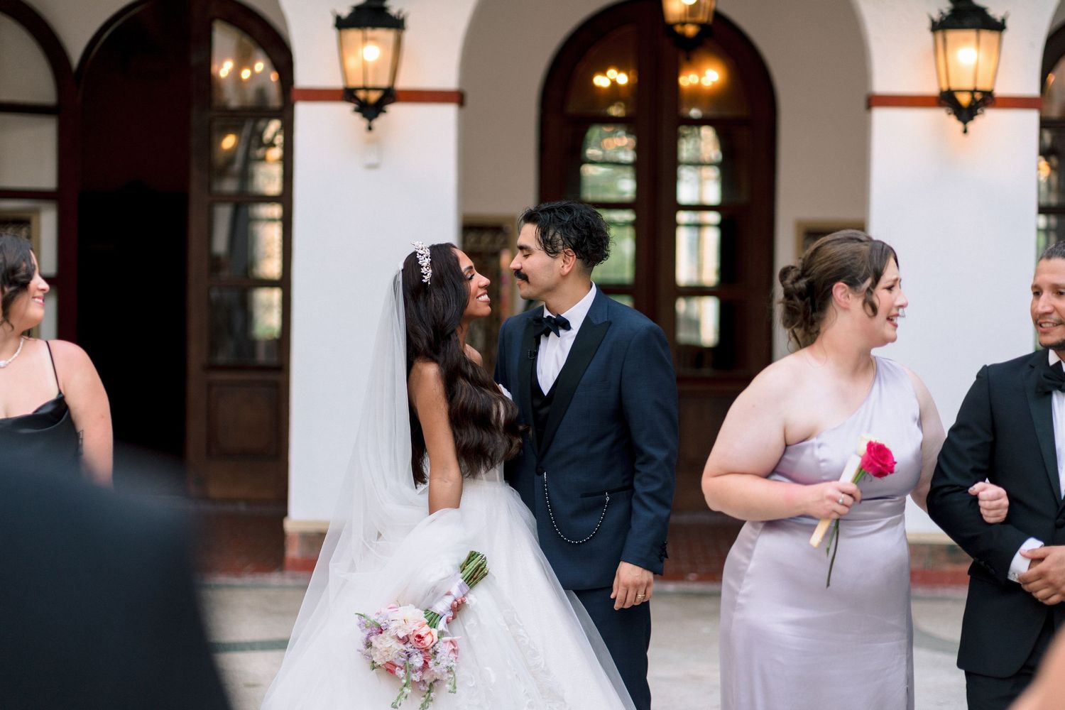 Smiling bride and groom share a kiss outside elegant venue surrounded by wedding party.