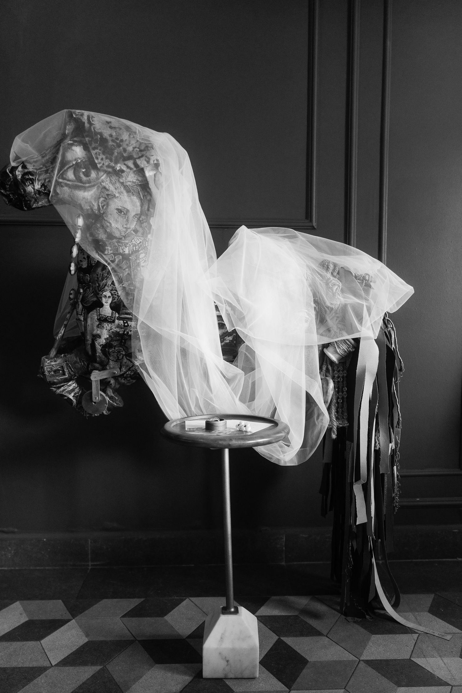 Black and white photo of a decorative horse sculpture draped with sheer fabric and ribbons on a pedestal stand.