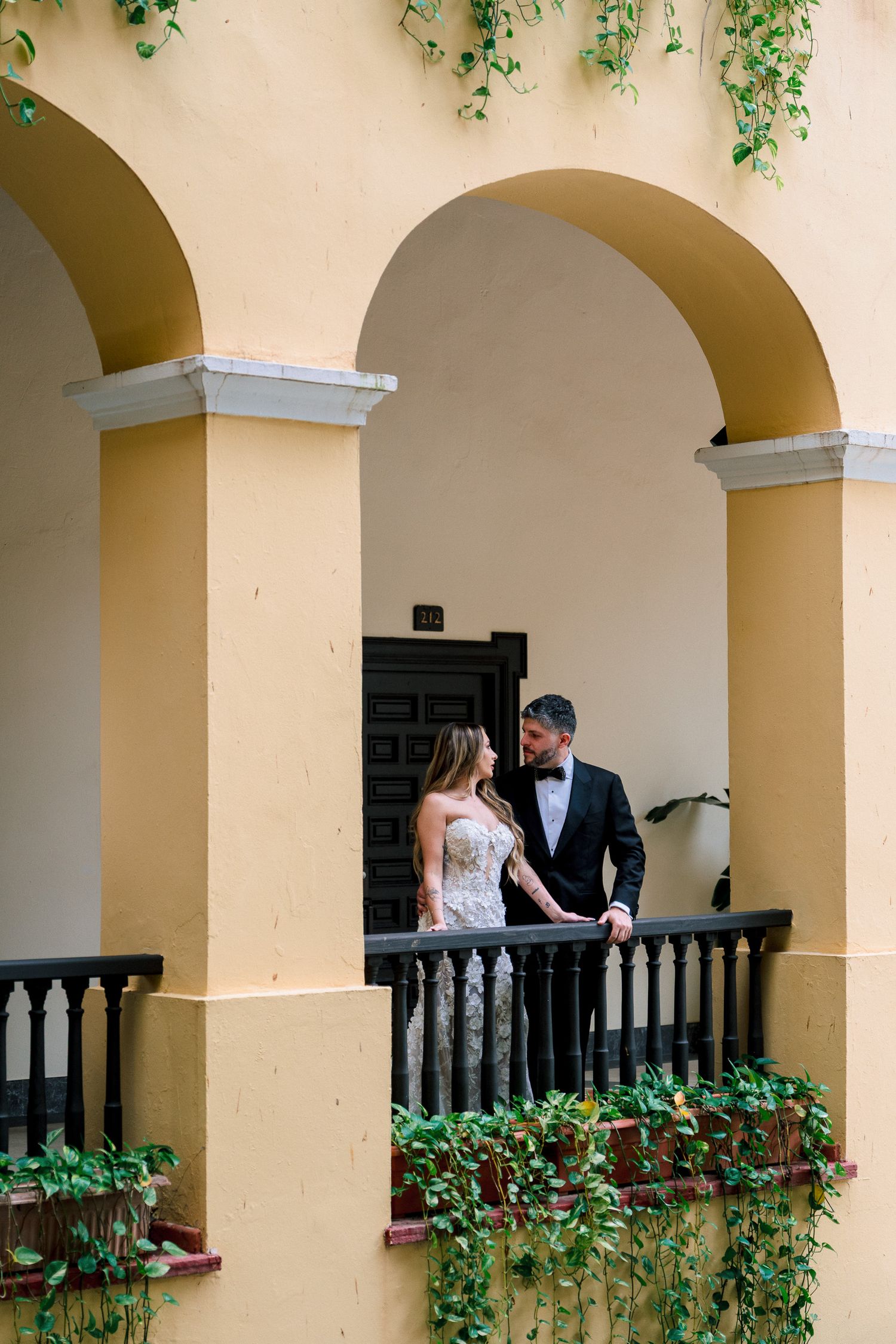 Bride and groom share a romantic moment on a yellow colonial balcony with arches and lush ivy in a historic setting.