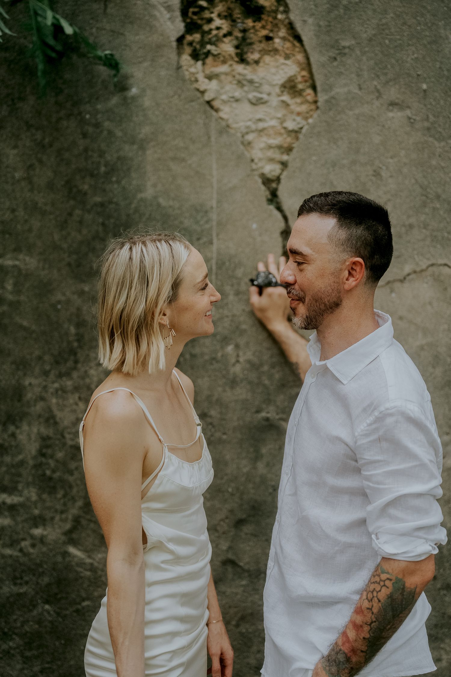 A smiling couple in white outfits face each other against a rustic stone wall, the man showing a tattoo sleeve.