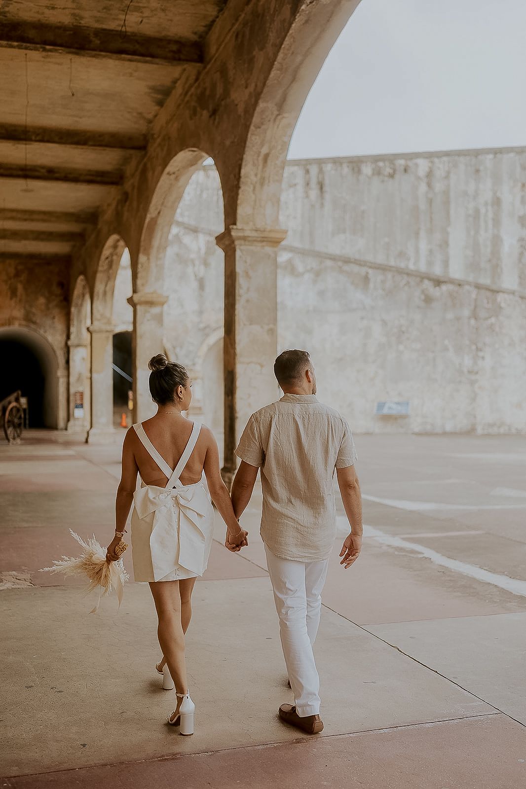 Couple in white outfits holding hands walking under stone arches at a historic venue.