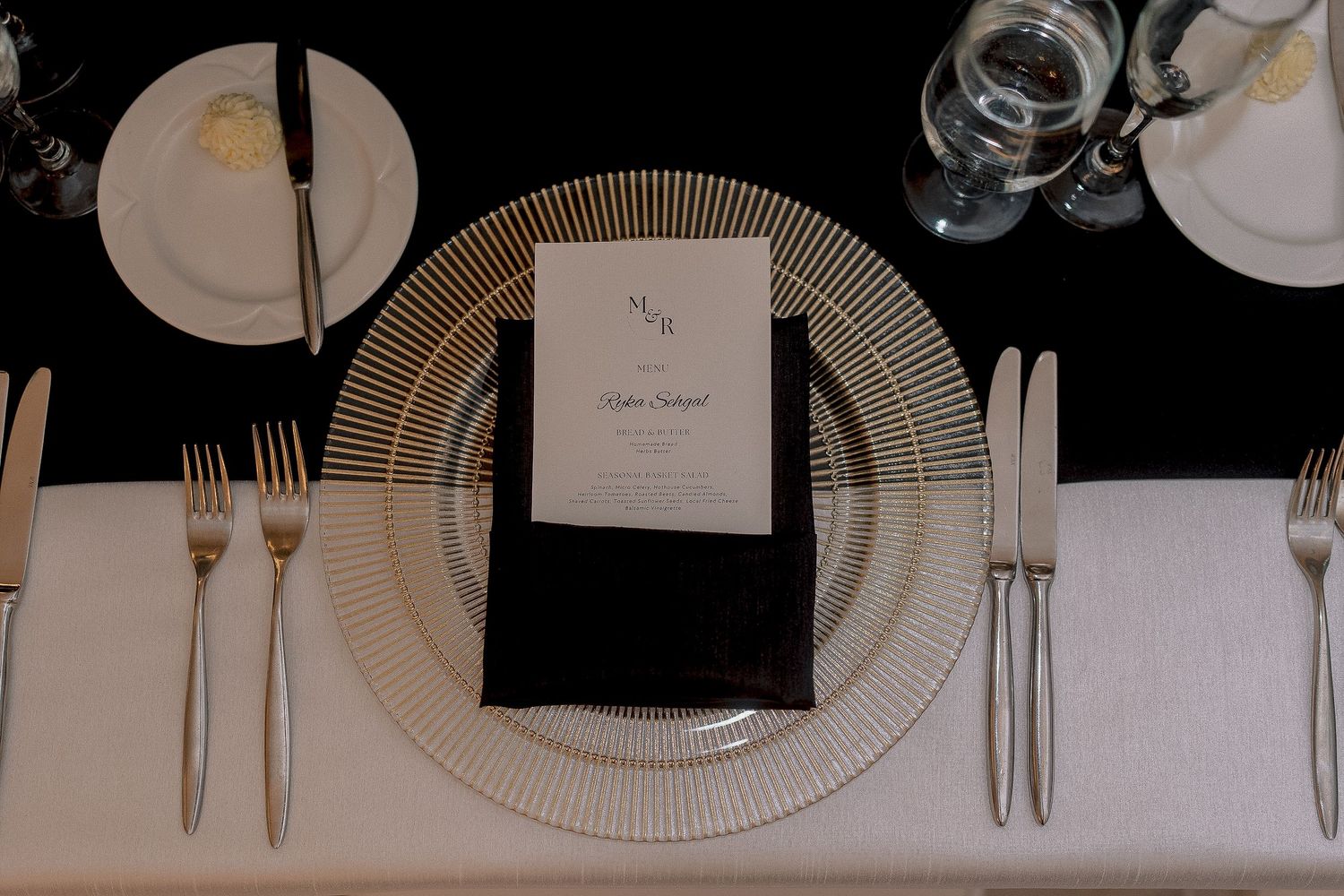 Elegant wedding reception place setting with gold charger plate, black menu card, and crystal glassware.