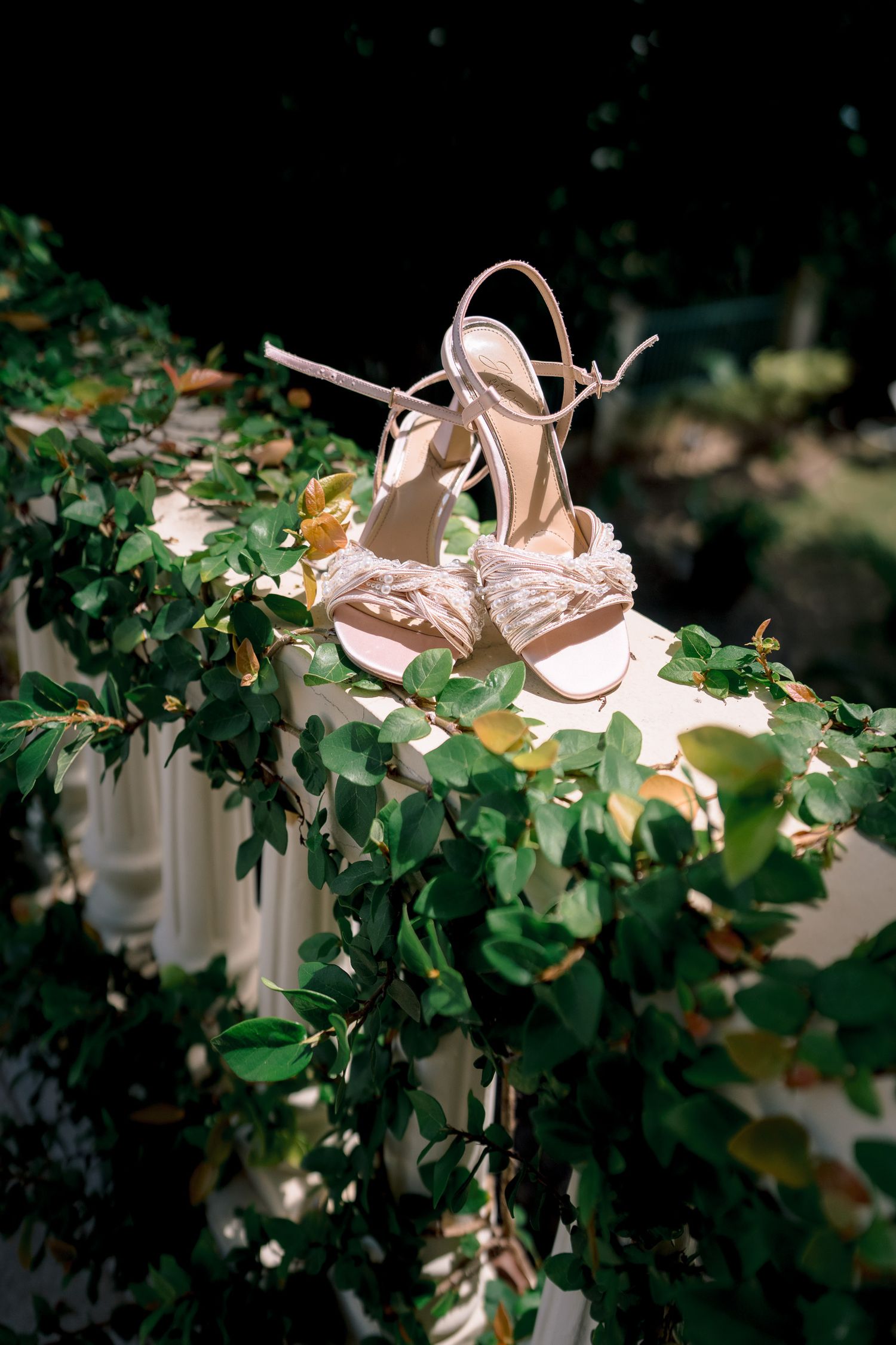 Elegant ivory bridal heels with feather details and ankle straps displayed on a white railing adorned with lush green ivy.