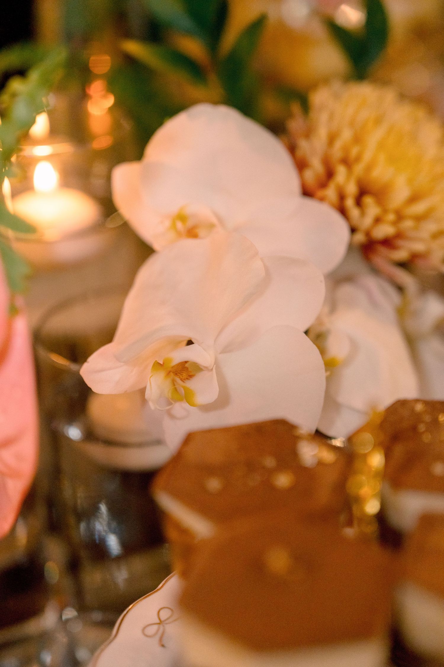 White orchids and yellow dahlias wedding centerpiece with glowing candles and gold-glazed pastries on elegant table.