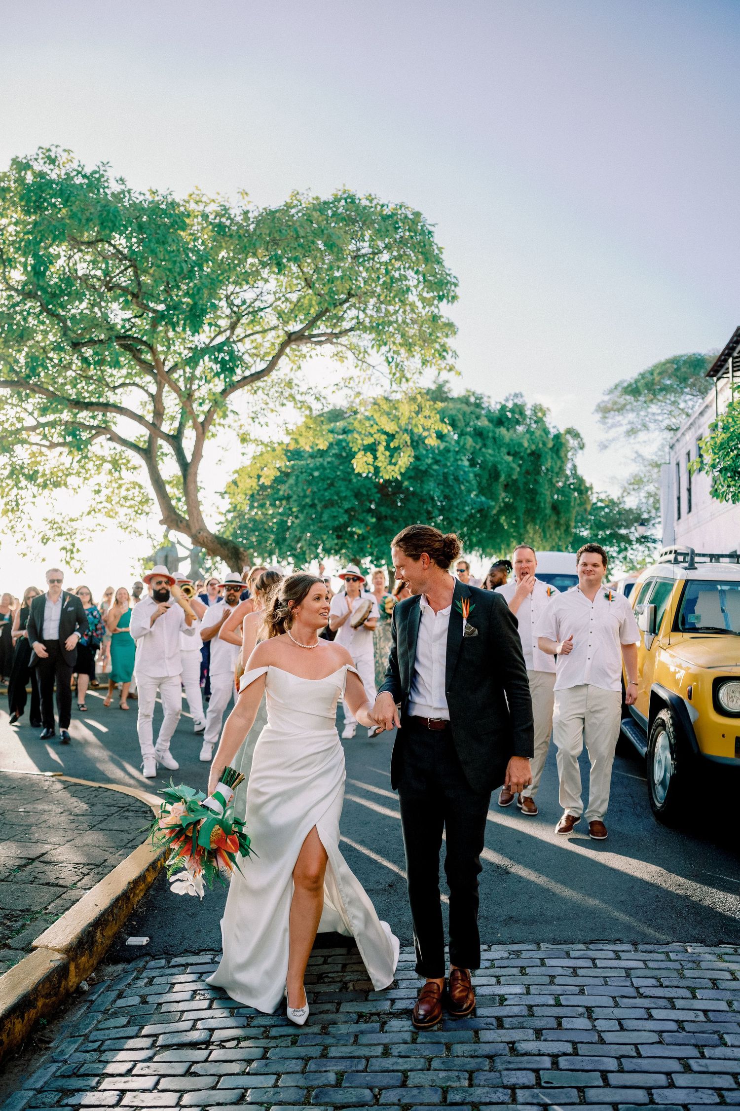 Bride and groom walk hand-in-hand on cobblestone street followed by wedding guests during outdoor celebration.
