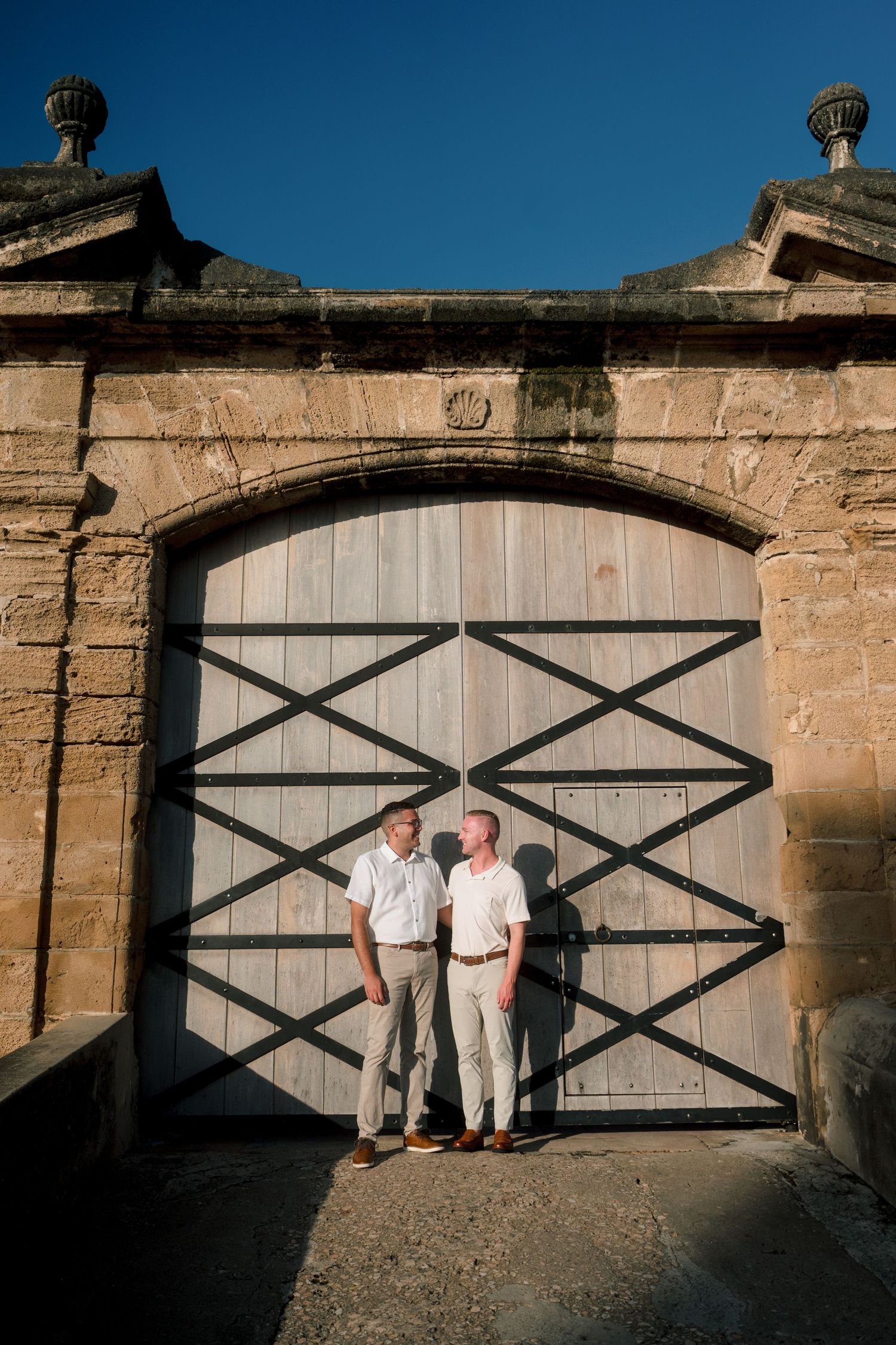 Two men in white shirts stand before a large wooden arched gate at a historic stone building.