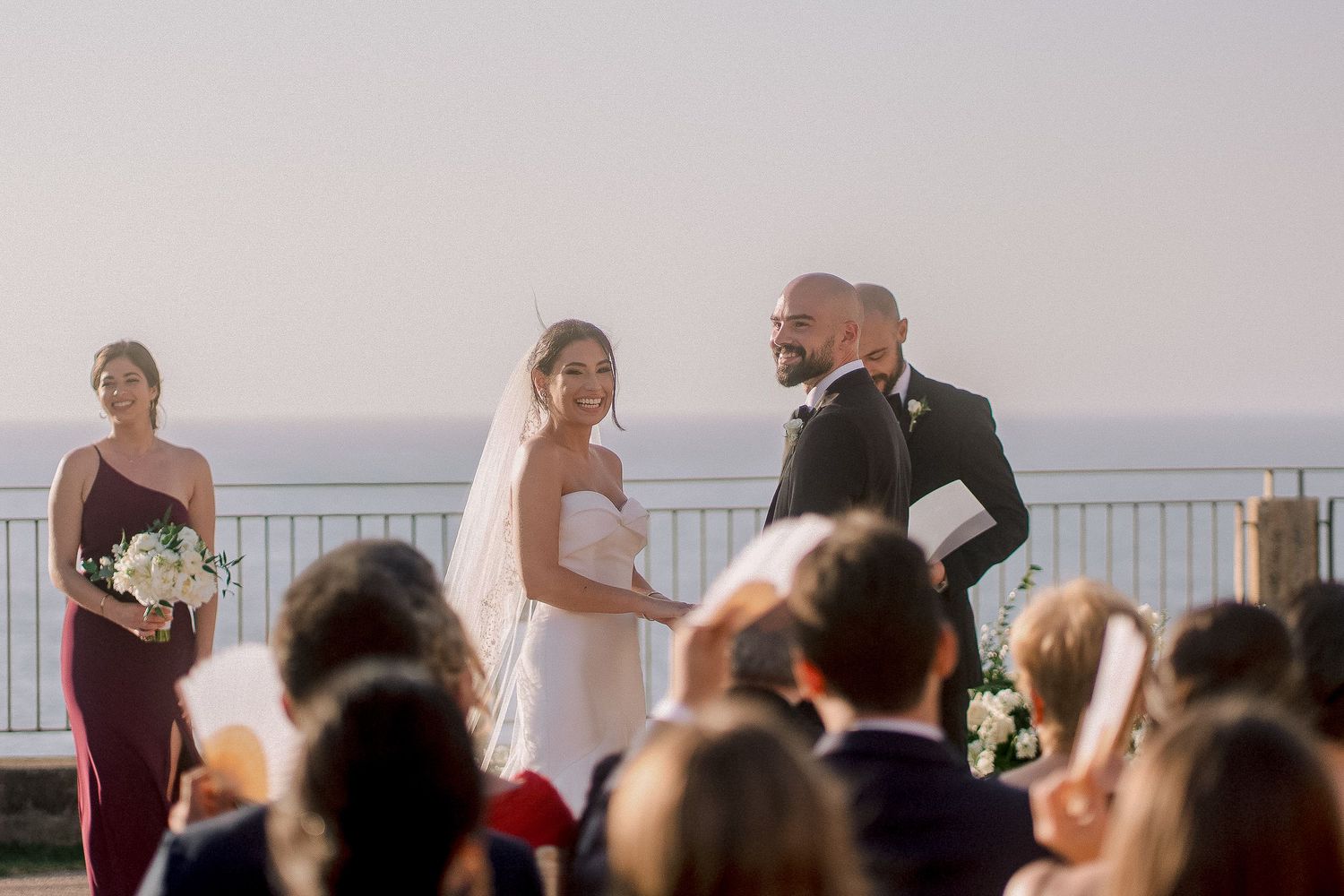 Bride and groom exchange vows at outdoor wedding ceremony with ocean view, bridesmaid in burgundy dress nearby.