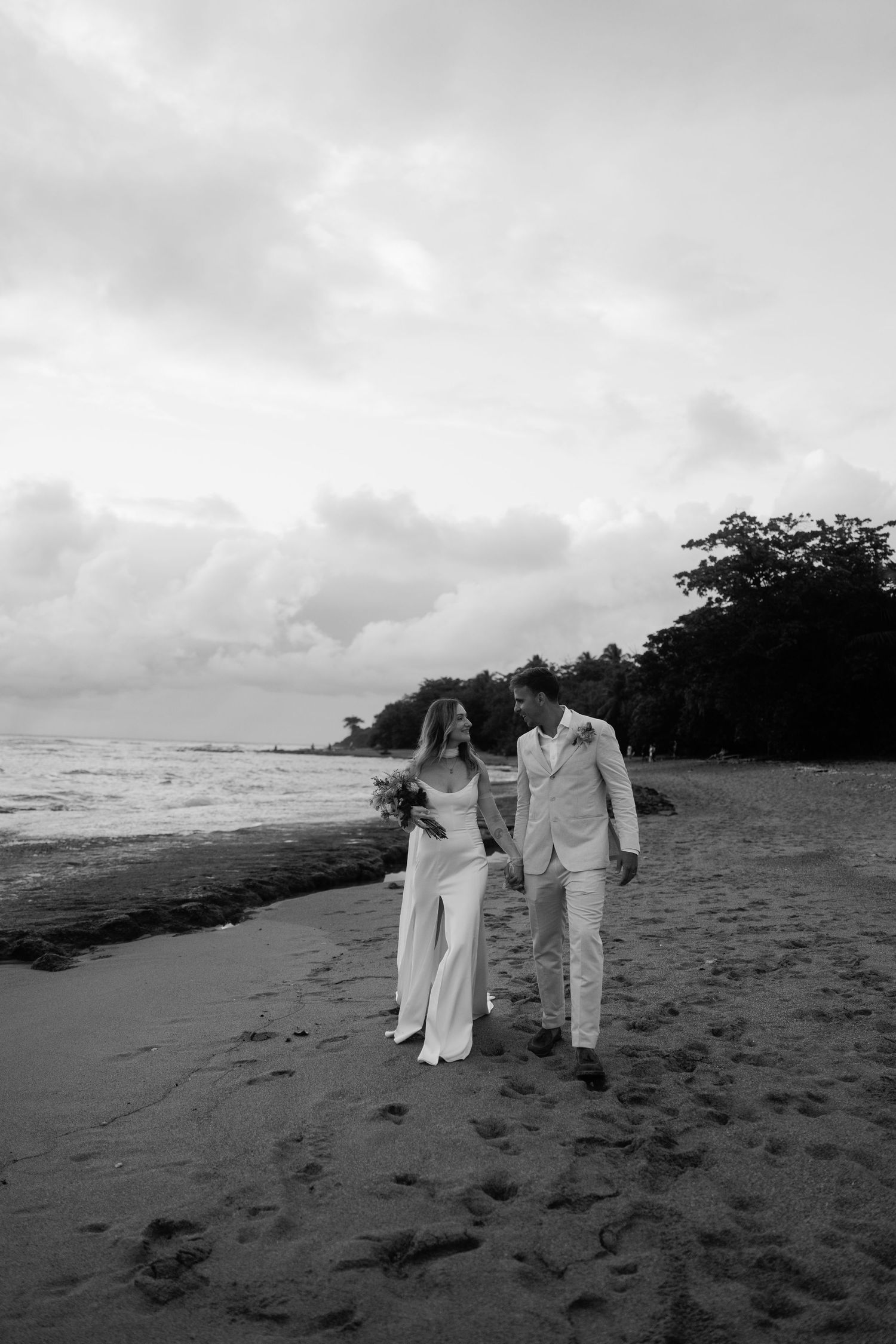 Black and white photo of a bride and groom walking hand in hand along a sandy beach with ocean waves and trees.