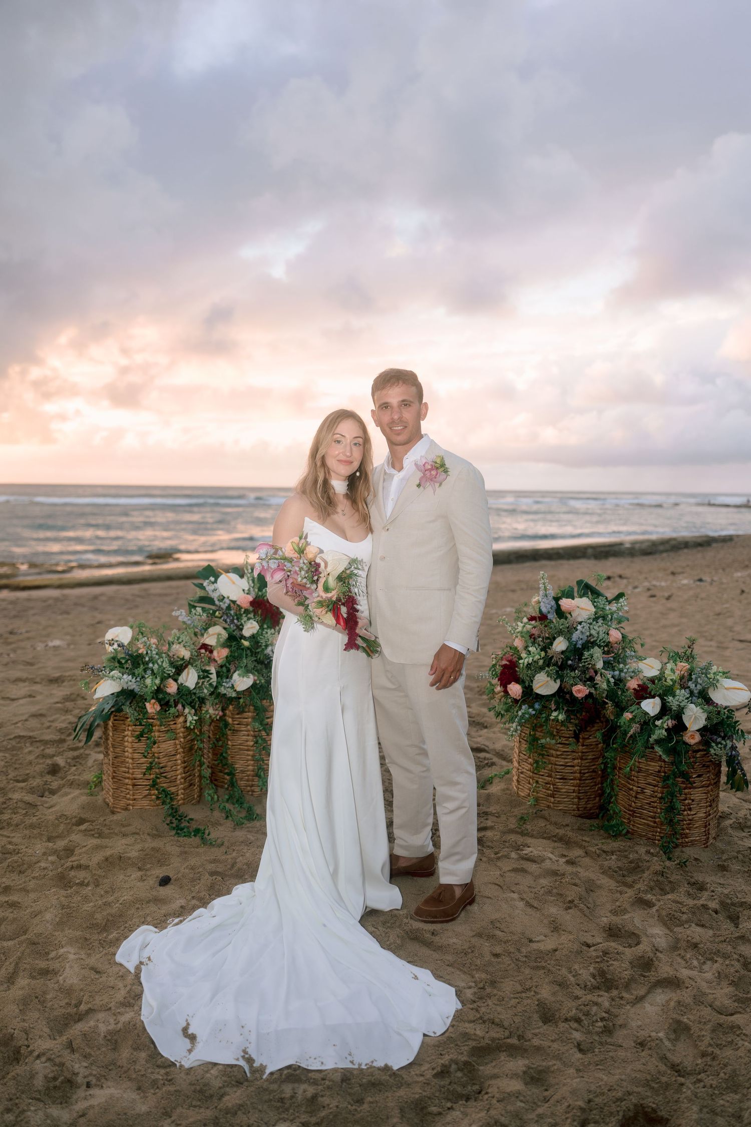 Bride and groom pose at beach wedding altar with floral arrangements and dramatic sunset sky.