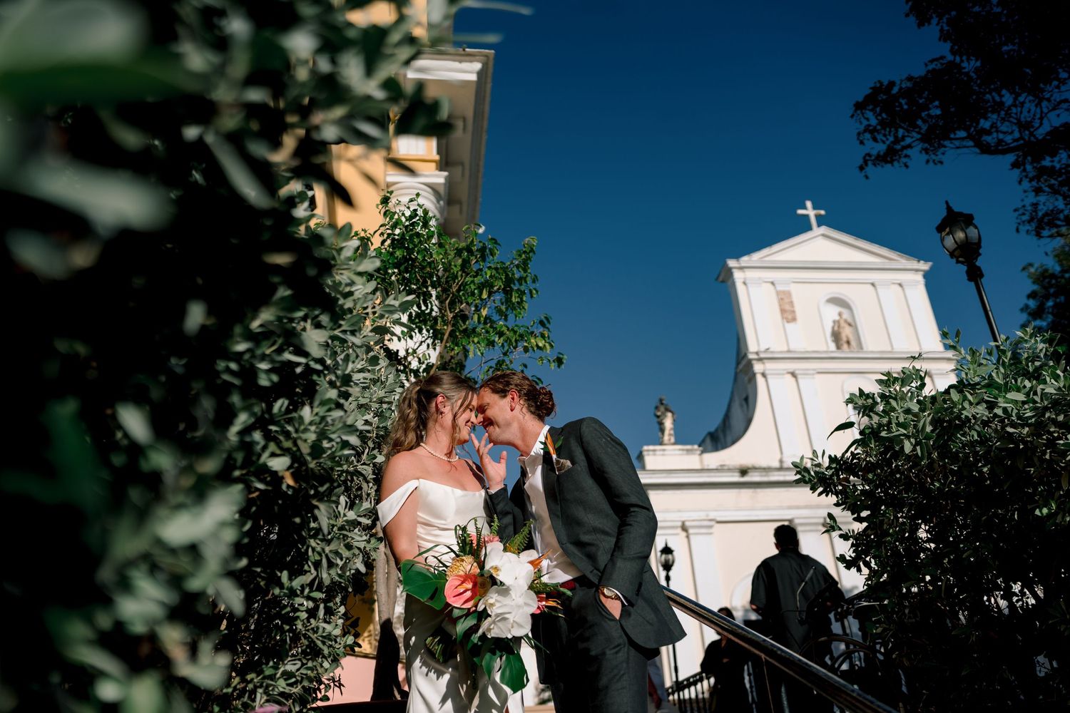 Bride and groom share a romantic moment outside a white church, surrounded by lush greenery on their wedding day.