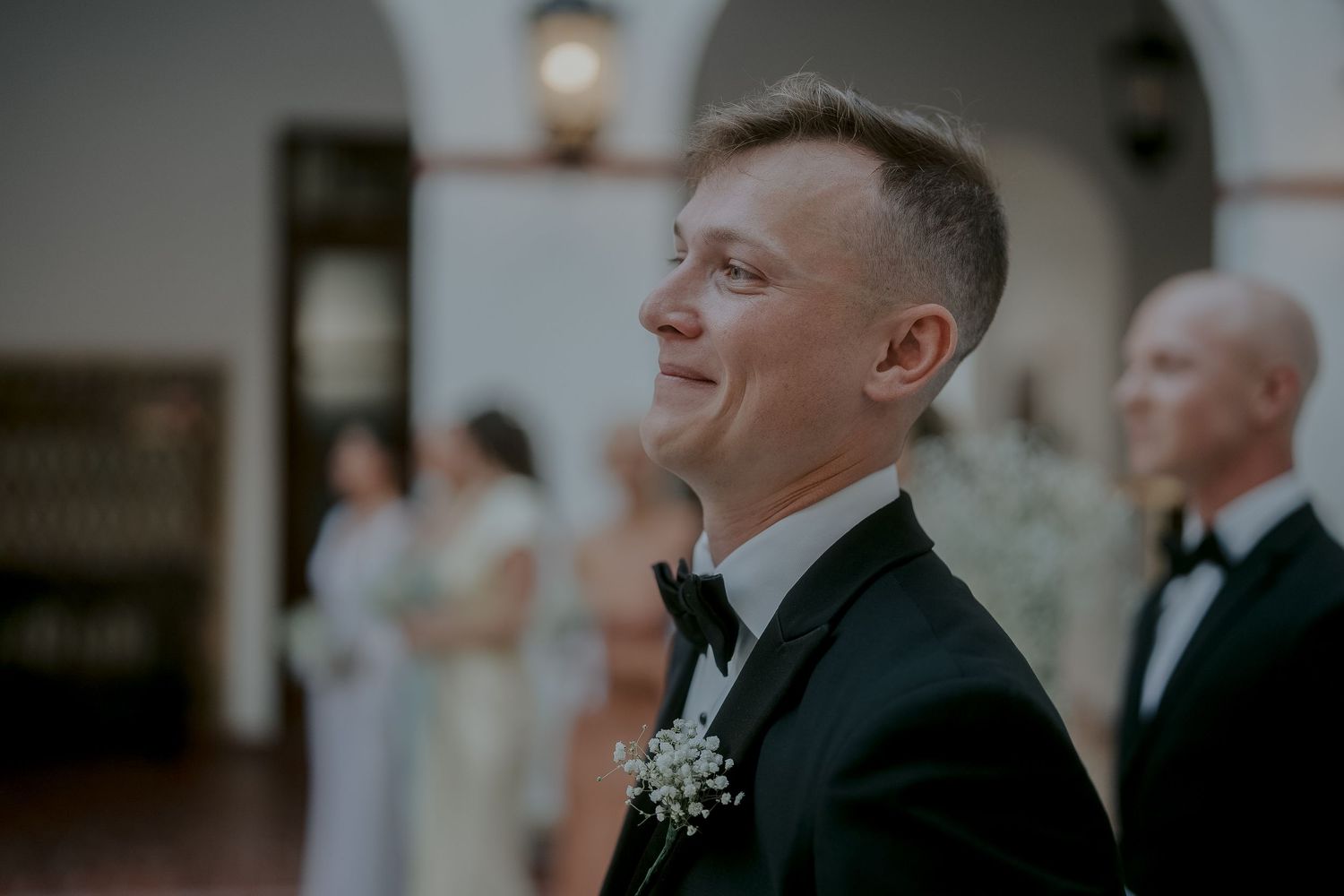 Groom in black tuxedo with boutonniere smiling joyfully at wedding ceremony.