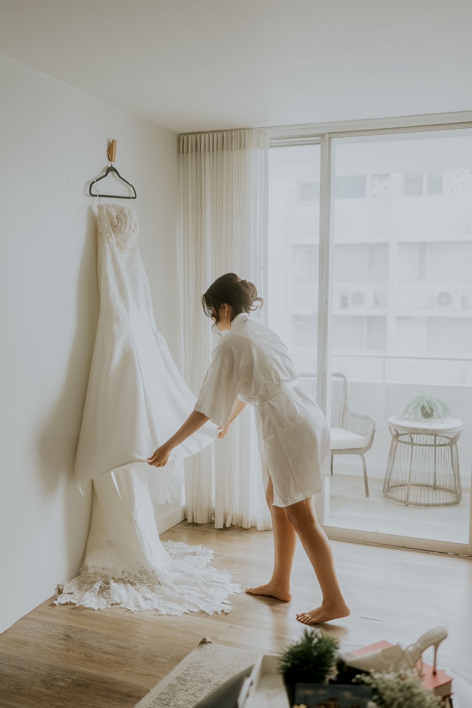 Bride in white robe arranging her wedding gown hanging by a sunlit window in a modern hotel room.