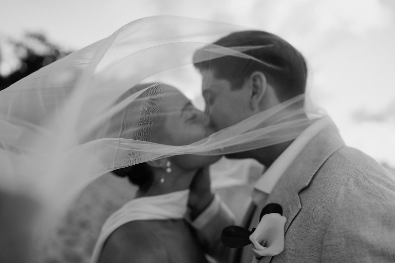 Bride and groom share tender kiss draped under flowing white veil in black and white photo.