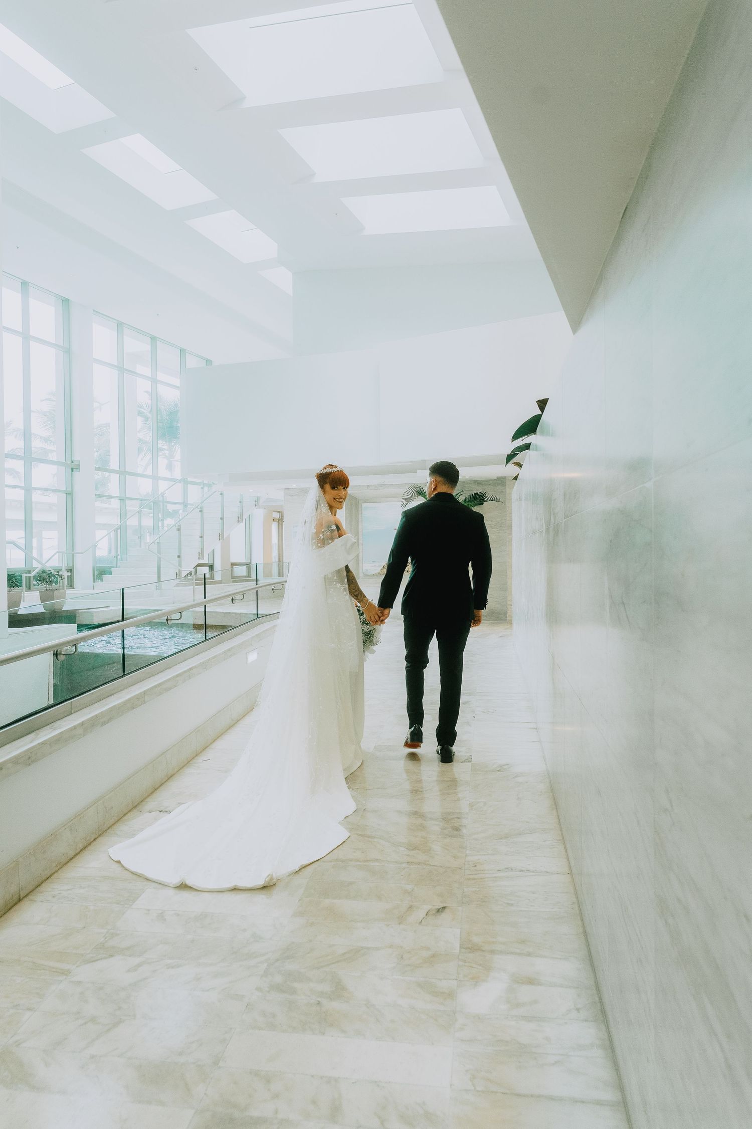 Bride in white gown and groom in black suit walk hand in hand through bright marble hall.