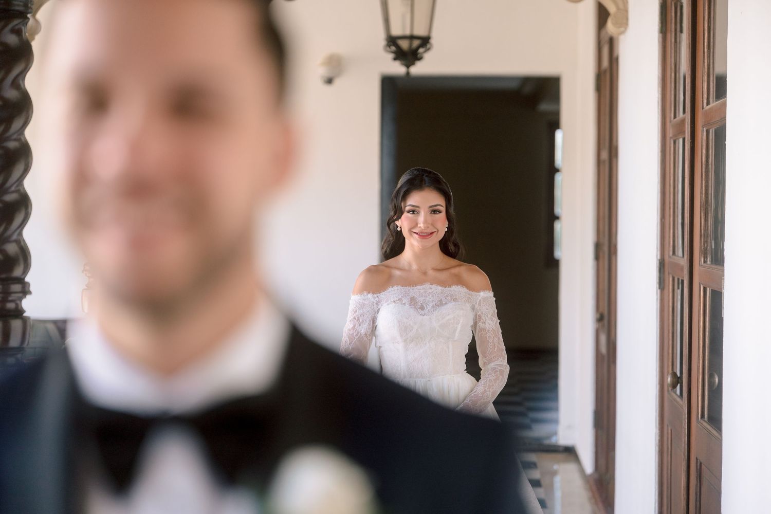 Bride in off-shoulder lace wedding gown smiling at groom during romantic first look at elegant venue.