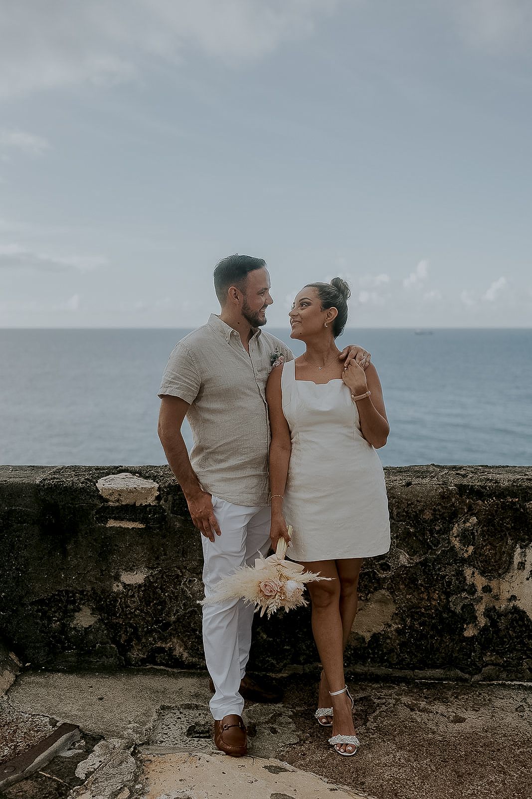 Couple in white attire sharing a romantic moment by a stone wall overlooking the ocean, bride holding a bouquet.