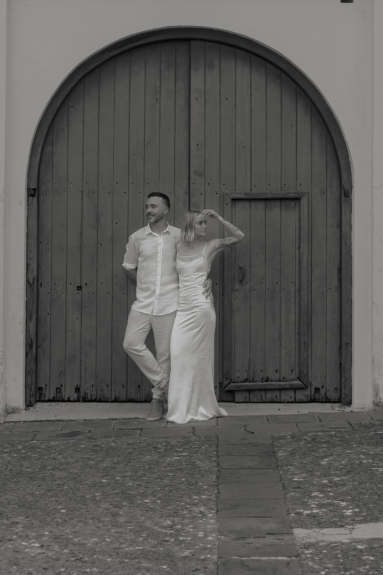 Elegant couple in white posing before a large arched wooden door in a black and white wedding photo.