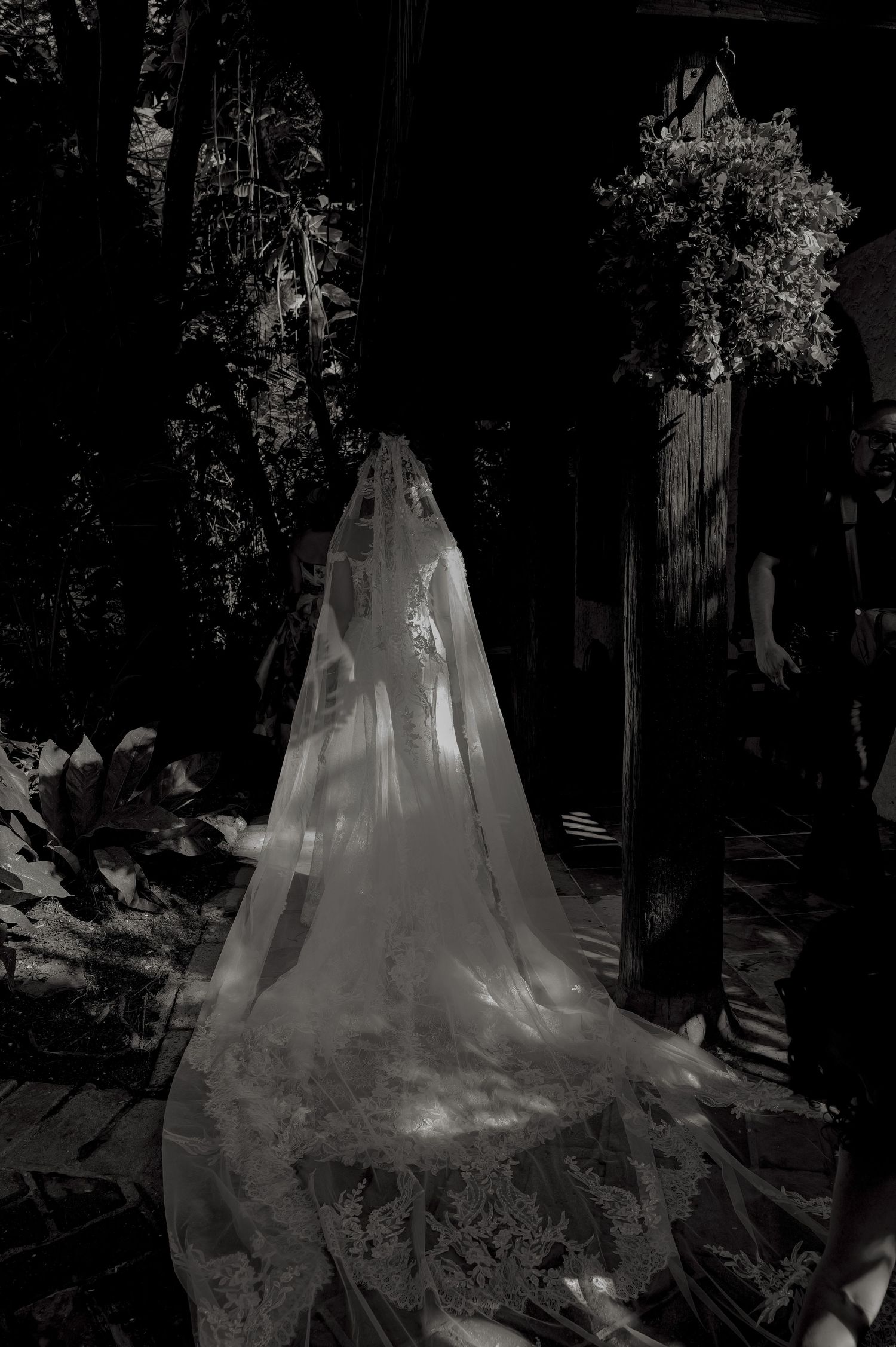 Black and white photo of bride in lace wedding gown and veil standing in a lush garden ceremony.