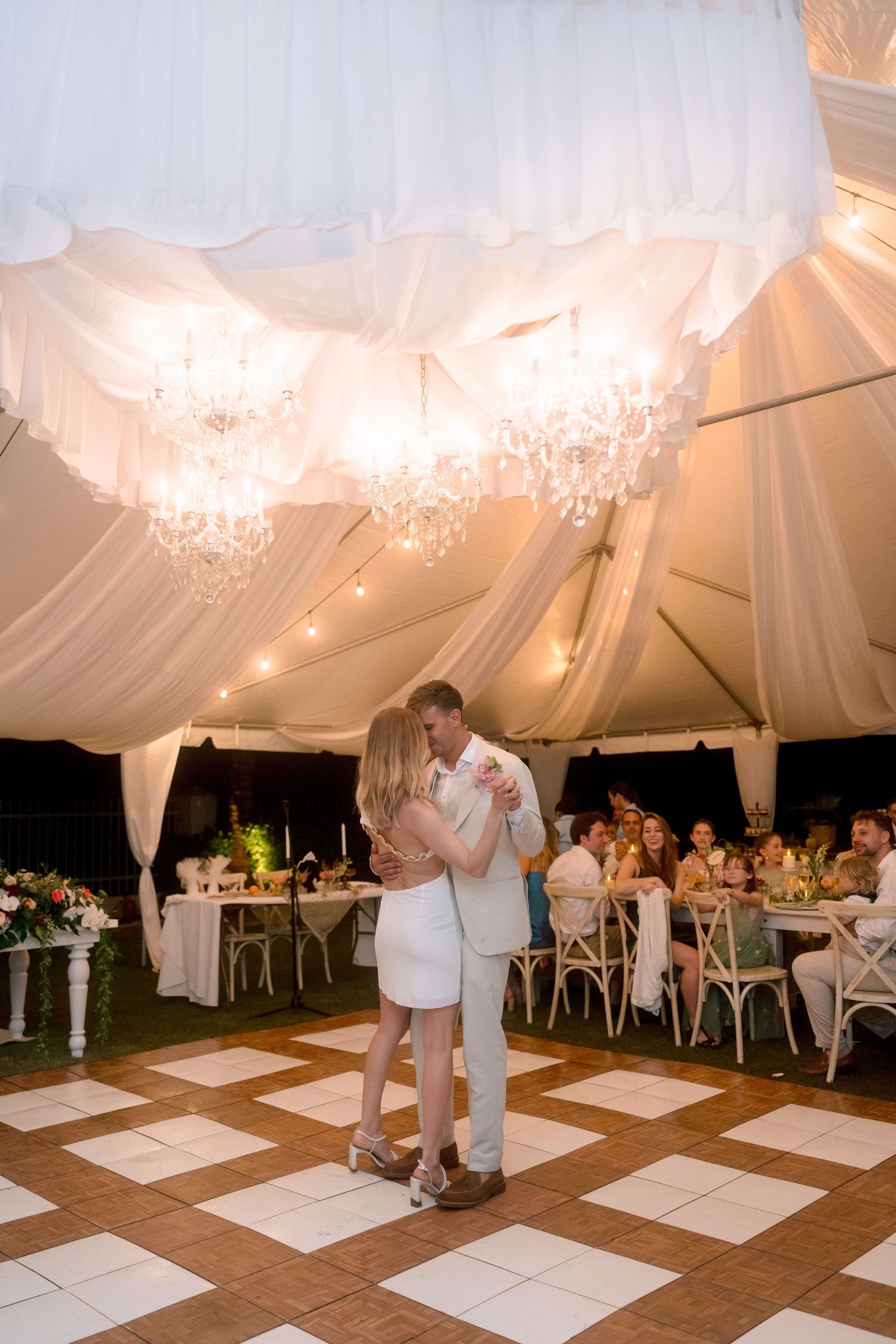 Newlyweds share first dance under crystal chandeliers in romantic white draped wedding reception tent.