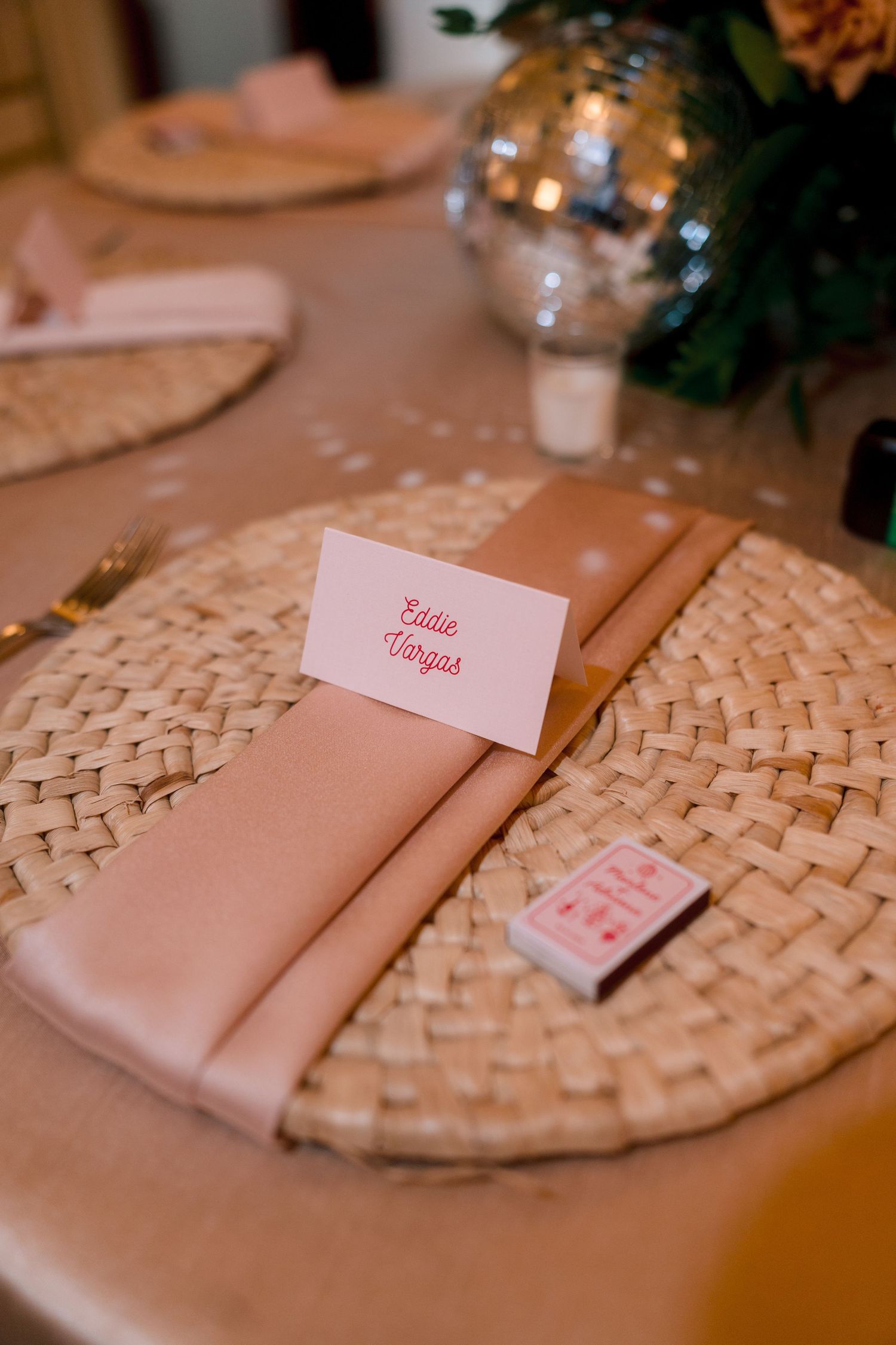 Elegant wedding place setting with pink napkin, woven charger, name card reading Eddie Vargas, and mini matchbox favor.
