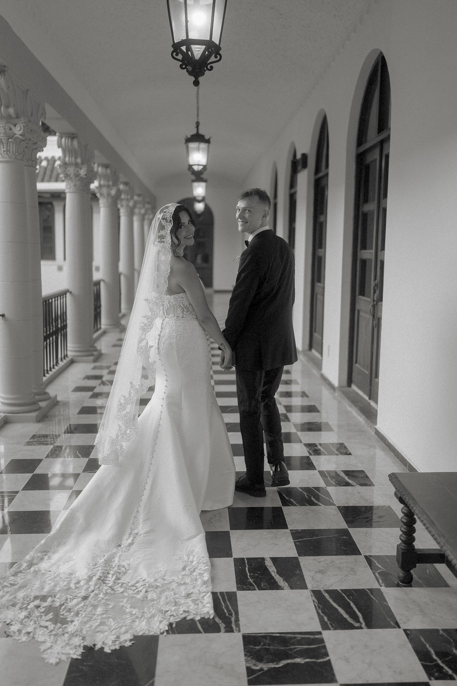 Black and white photo of bride and groom holding hands in ornate columned hallway with checkered floor.