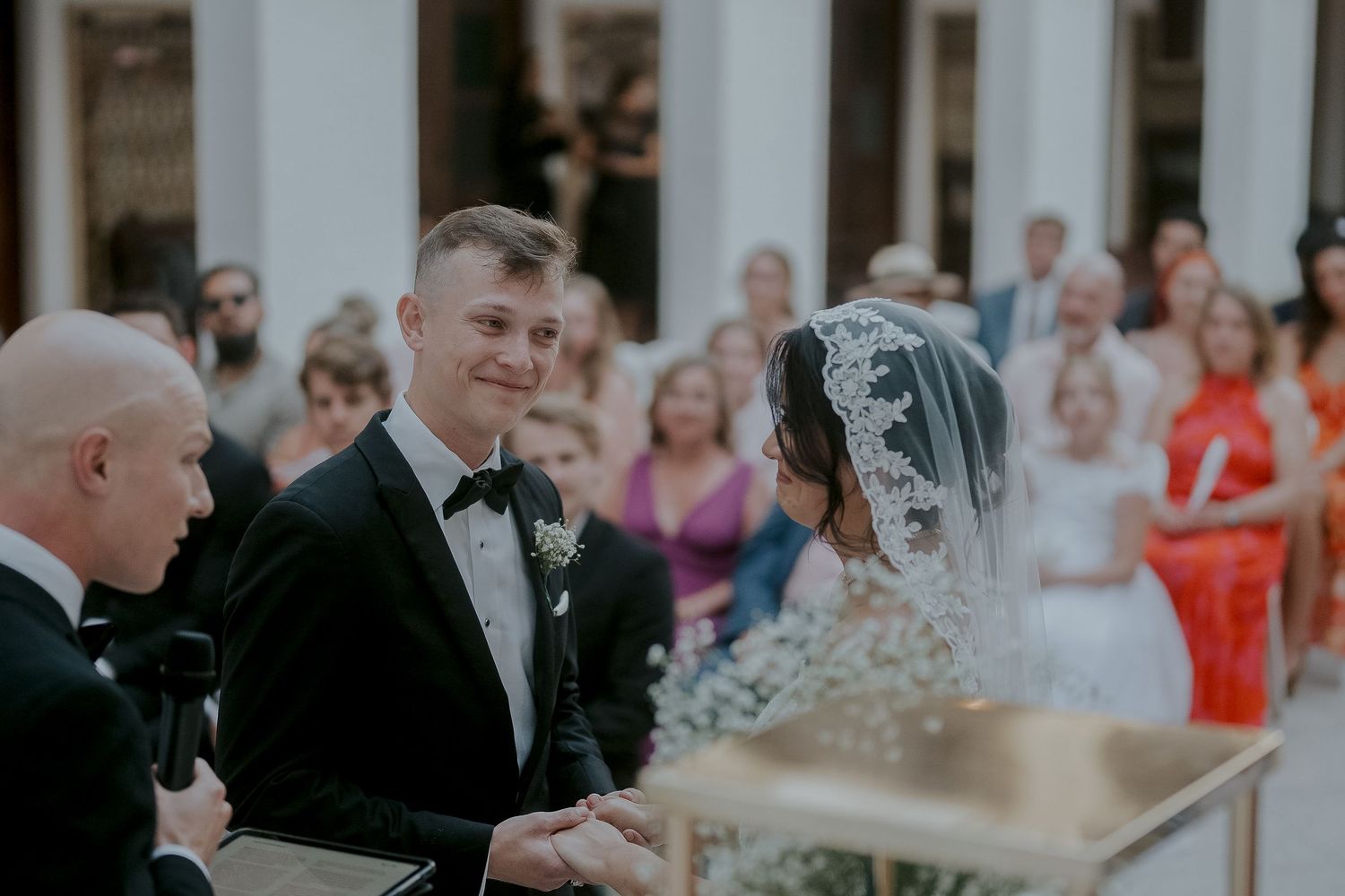 Groom in black tuxedo smiling at bride in lace veil during outdoor wedding ceremony with guests in background.