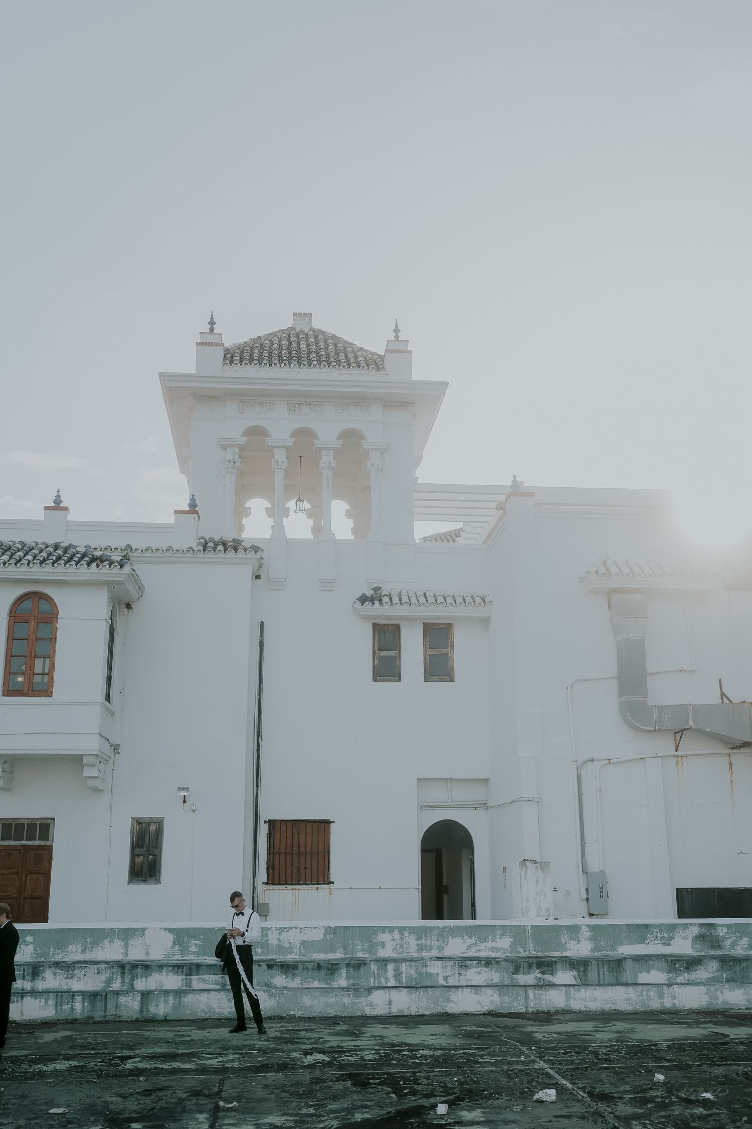 A person stands before a grand white Spanish-style building shrouded in mist on an overcast day.