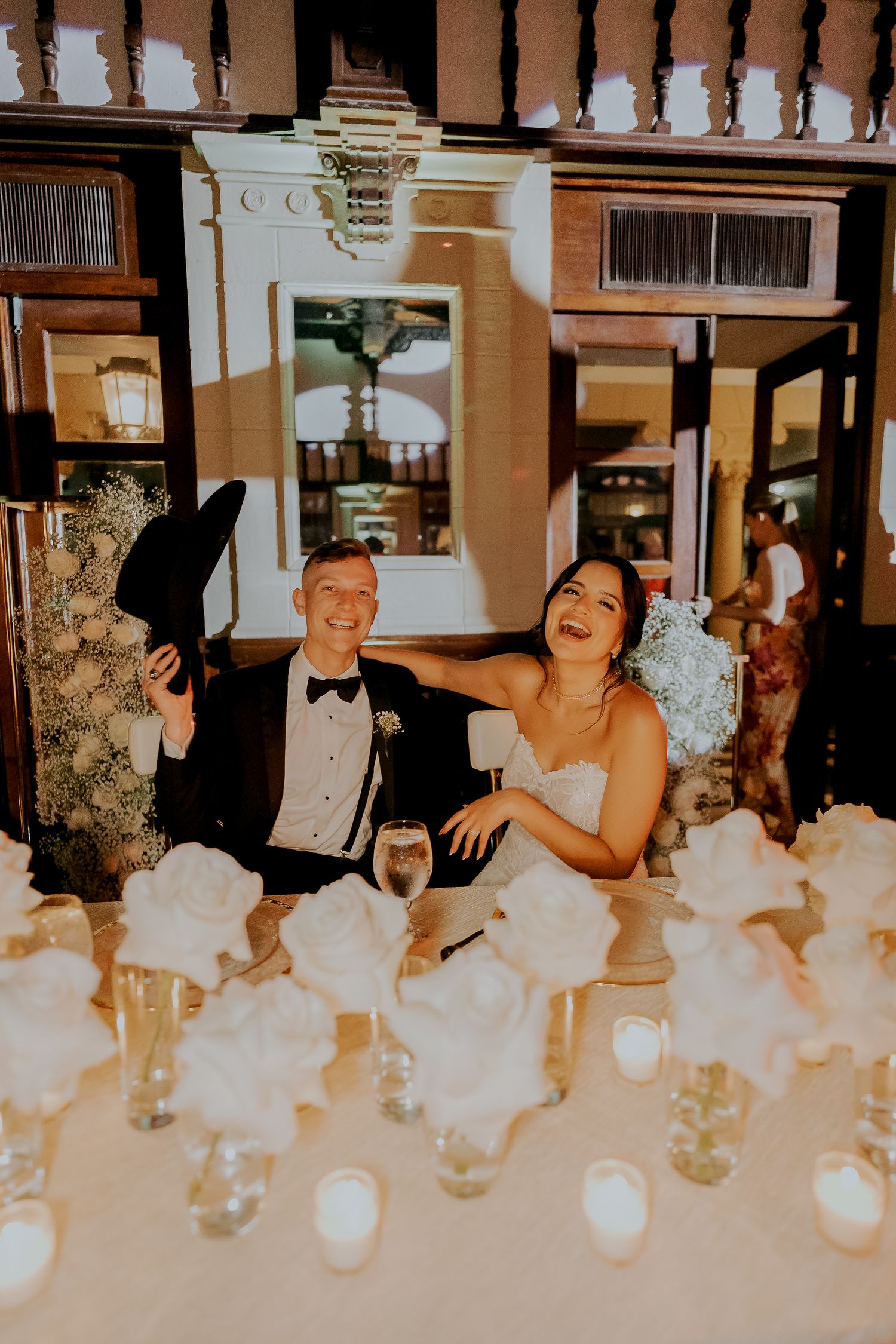 Laughing bride and groom sit at sweetheart table surrounded by white roses and candles at wedding reception.