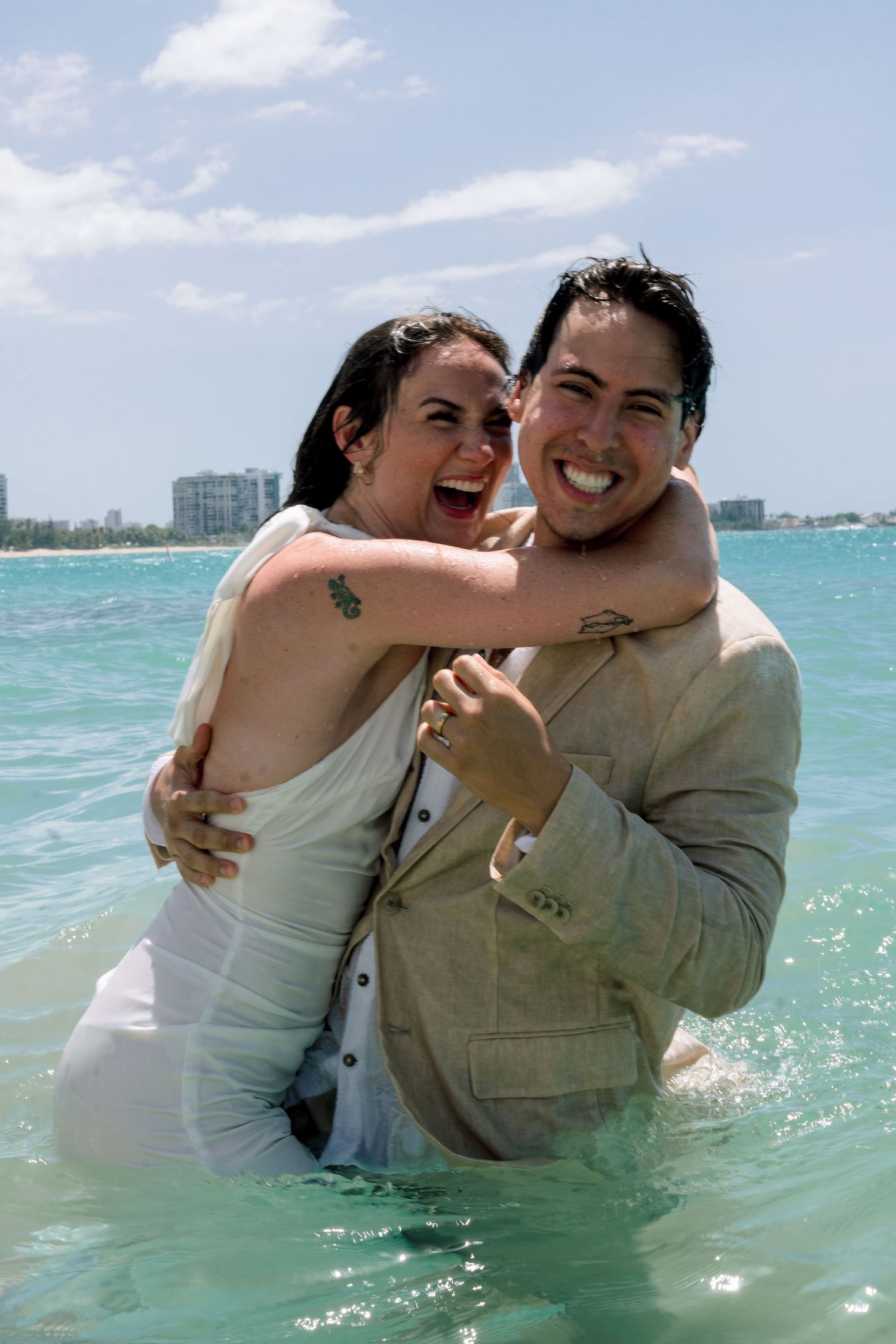 Happy couple in wedding attire laughing and embracing in turquoise ocean water on a sunny beach day.