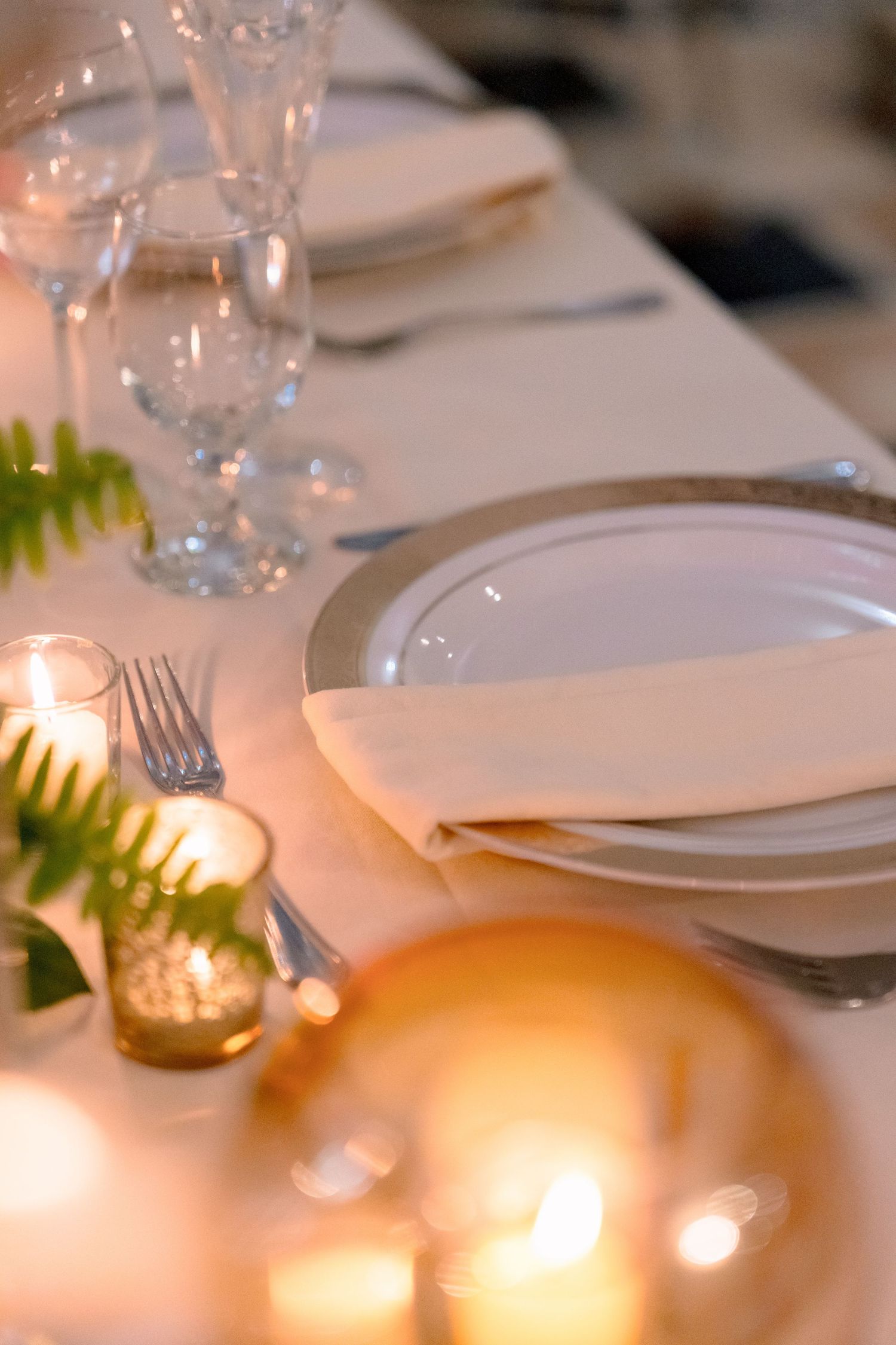 Elegant wedding table setting with white plates, crystal glasses, ferns, and glowing candles.