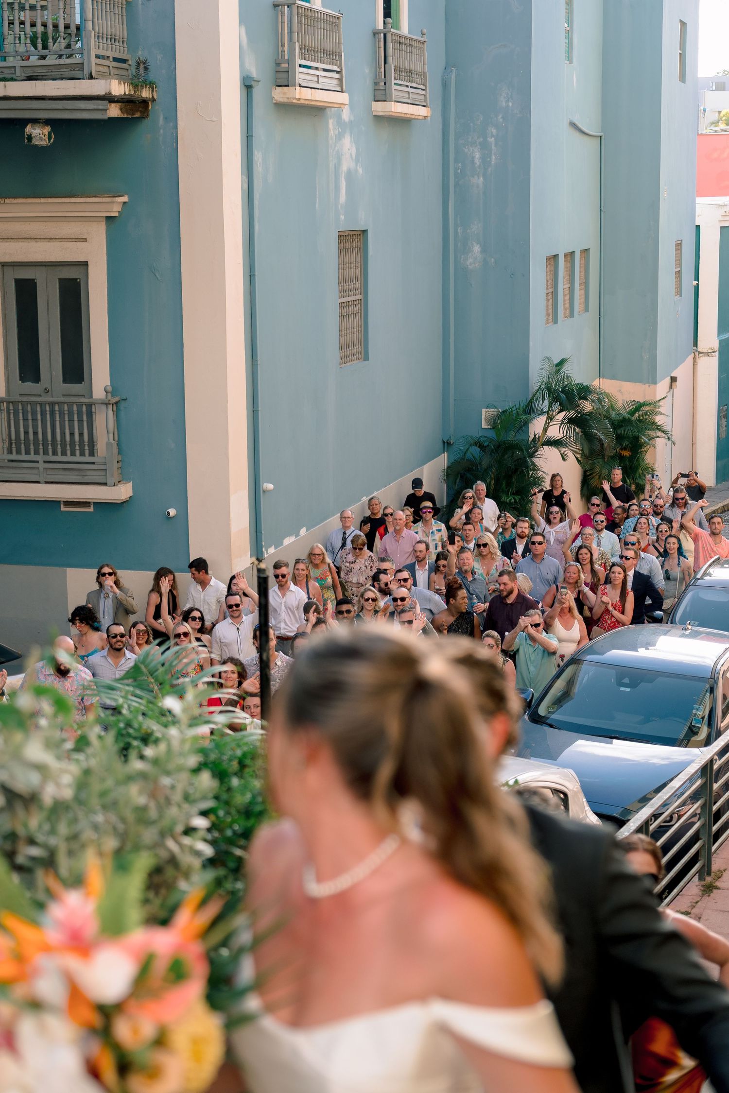 Bride and groom on balcony overlooking cheering wedding guests gathered on a vibrant blue street below.