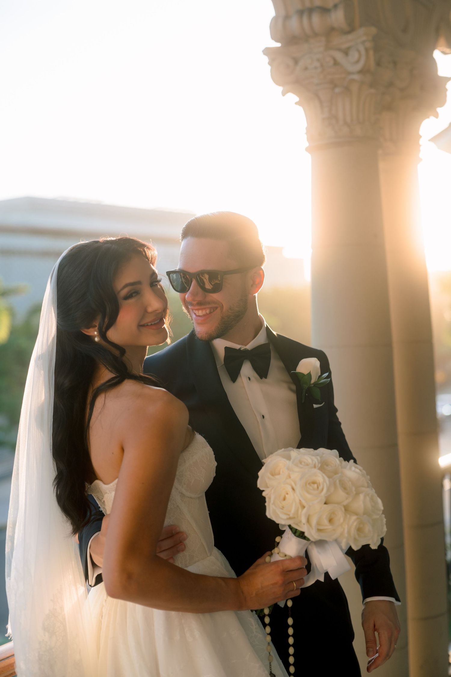 Smiling bride and groom in tuxedo holding white rose bouquet, posing in golden sunset light near ornate column.
