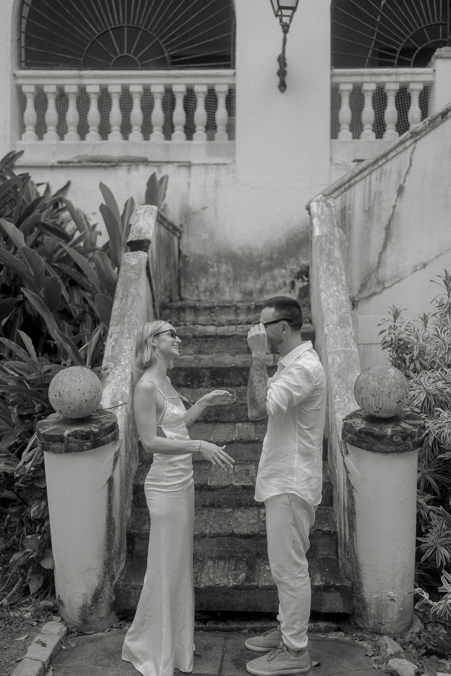 Black and white photo of a couple in elegant white attire standing by vintage stone steps surrounded by tropical plants.