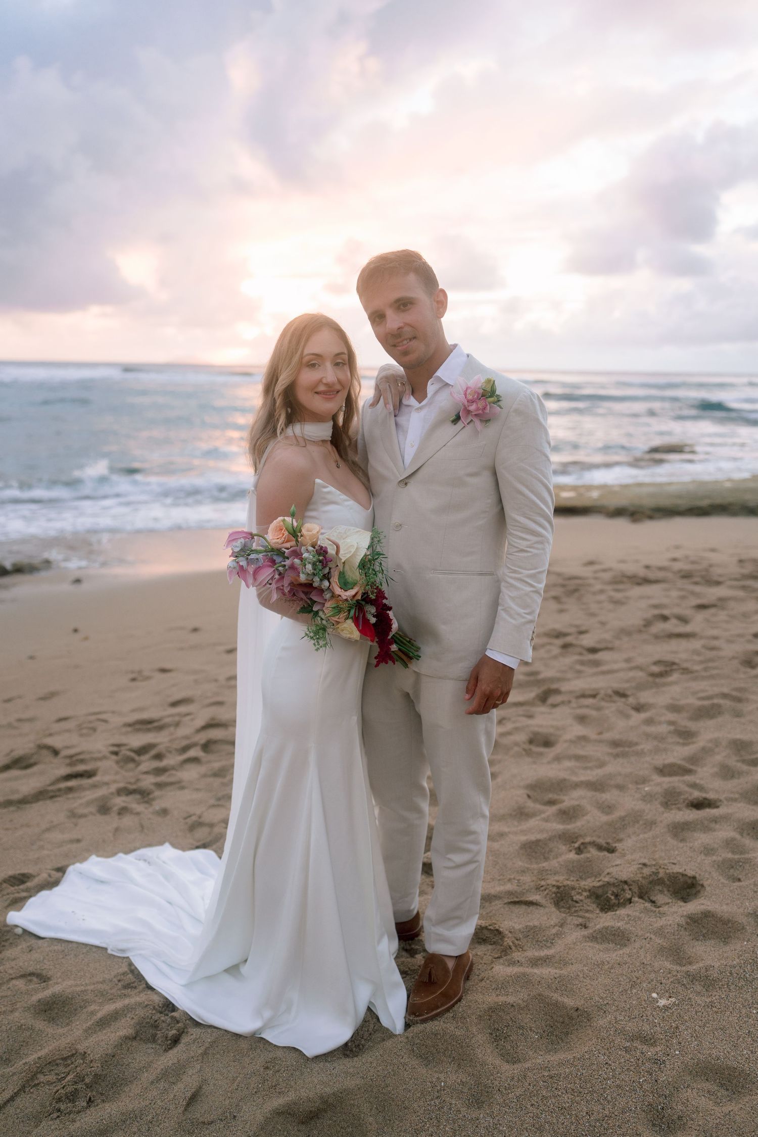 Smiling bride and groom pose together on sandy beach holding colorful bouquet during golden sunset.