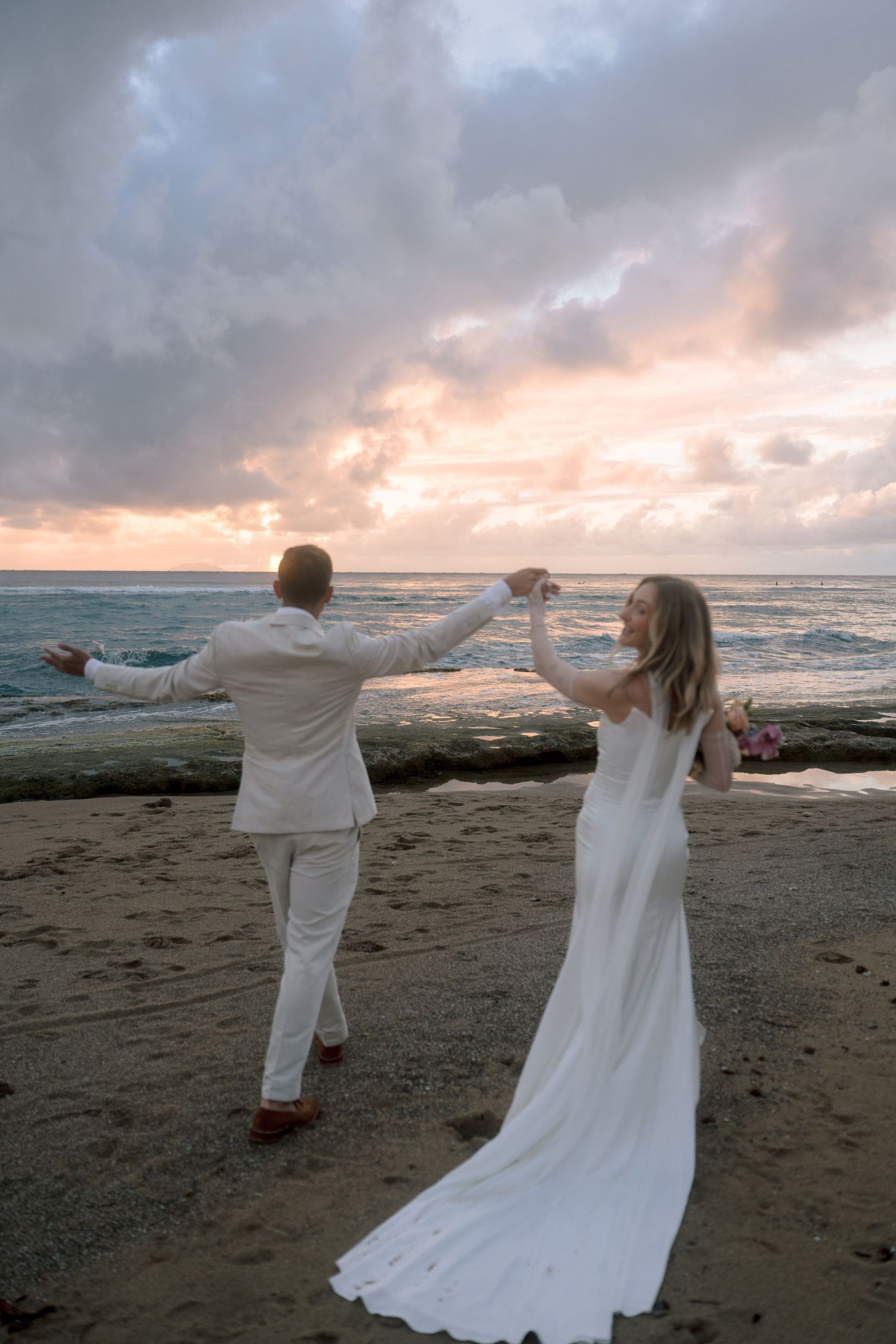 Bride and groom dancing joyfully on sandy beach at sunset, waves crashing behind them in colorful twilight sky.