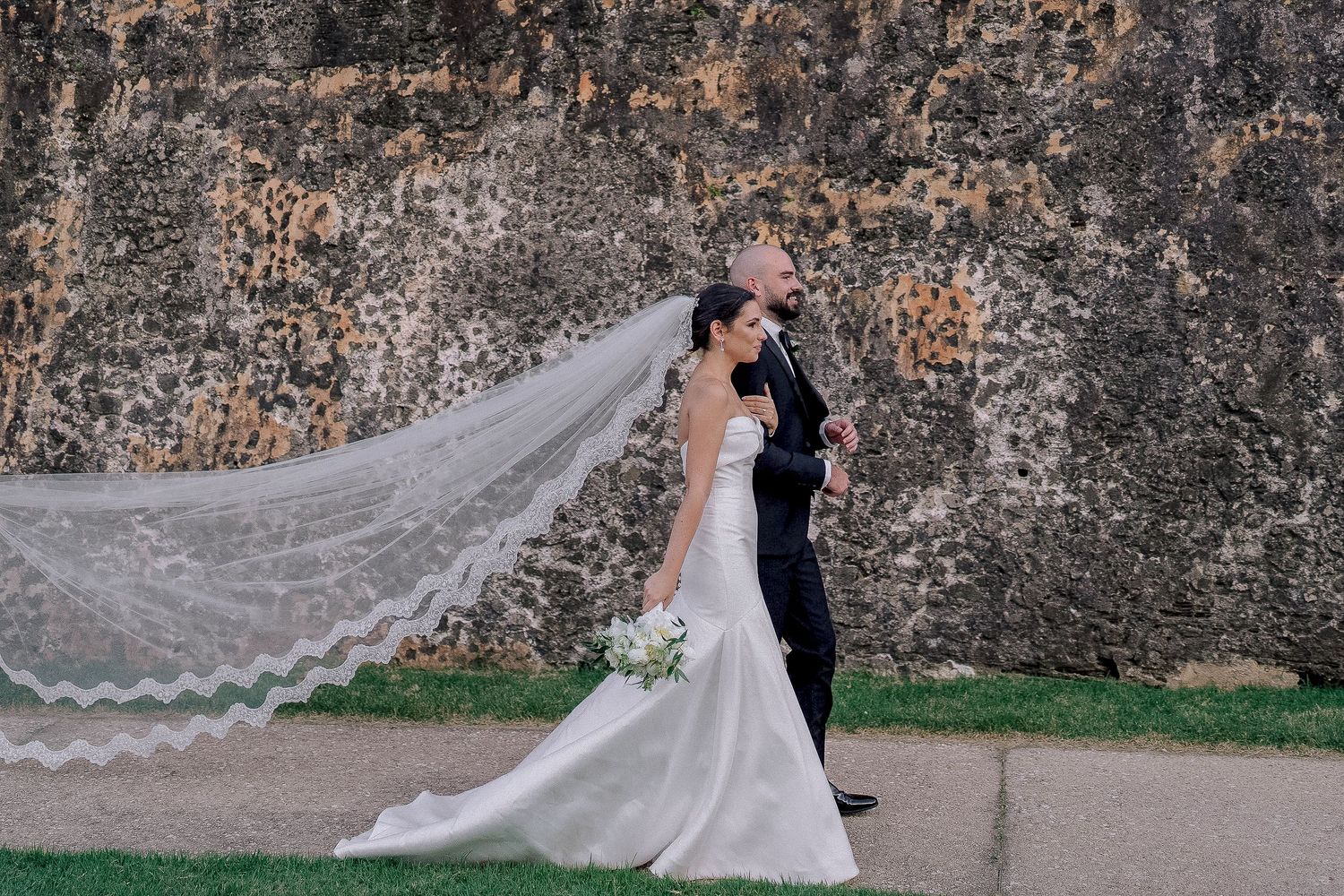 Bride and groom walk past ancient stone wall, her lace veil flowing dramatically in the wind.