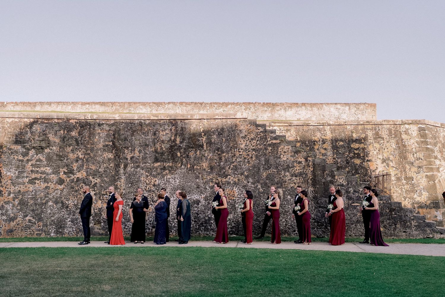 Wedding party in formal attire lined up against an ancient stone wall outdoors at dusk.