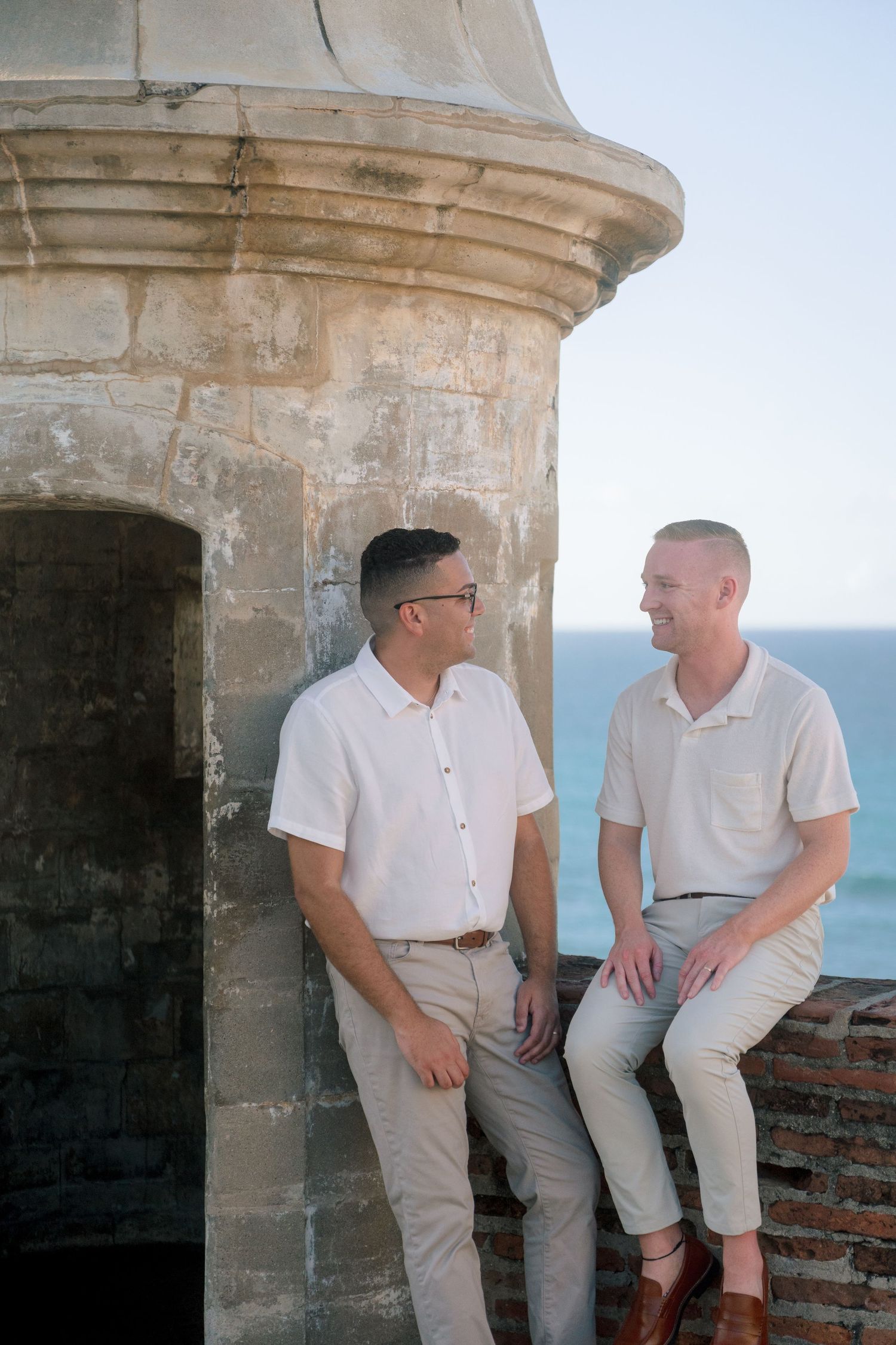 Two men in white shirts and khaki pants sit and stand by a historic stone turret overlooking the ocean.