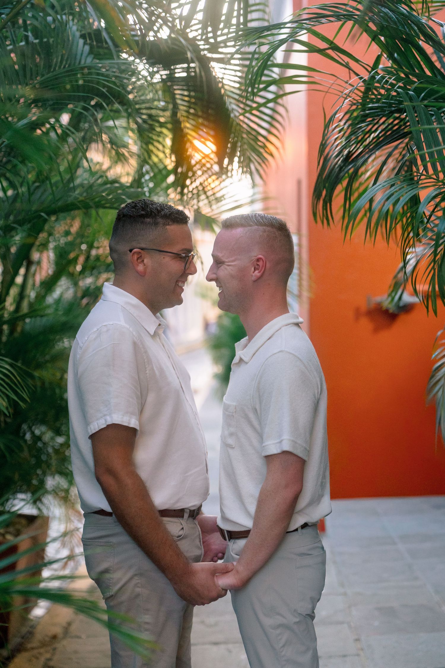 Two men in white shirts hold hands and gaze at each other among tropical palms near an orange wall.